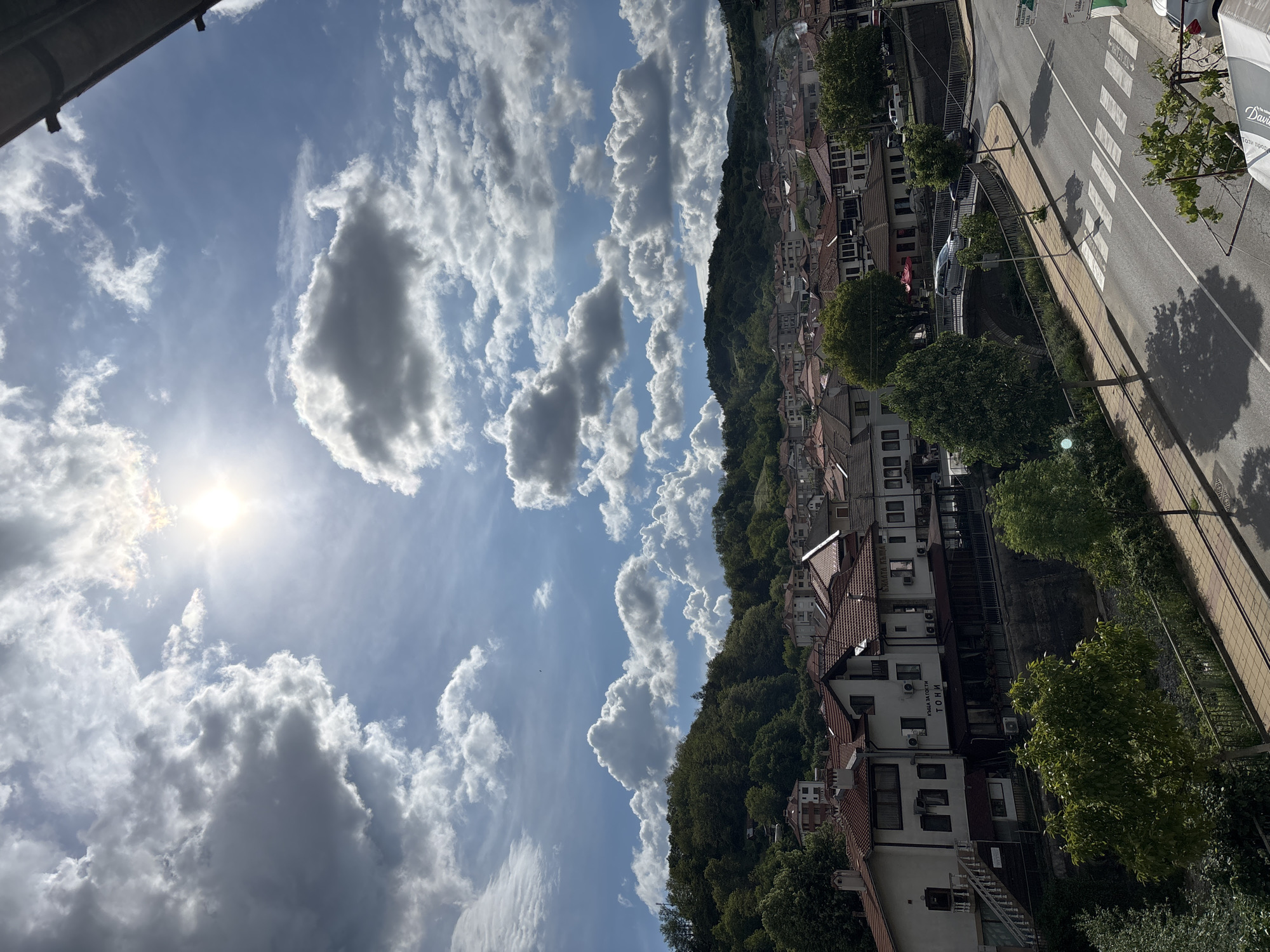 The image shows an outdoor view of Zlatograd, Bulgaria. It is a sunny day with a sky full of clouds. In the foreground, there are buildings with terracotta roofs nestled among trees. A road runs parallel to the buildings, with a pedestrian crossing visible. A small river runs parallel to the road. The background features a mountain covered in dense forest.