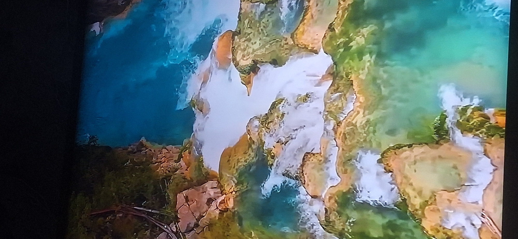 The image depicts a vibrant natural landscape with cascading waterfalls and lush greenery. The water is a striking turquoise blue, indicating clarity and possibly mineral content. Rocky formations, partially covered in moss and vegetation, frame the waterways. The scene appears to be an outdoor environment, possibly a park or natural reserve, with varying shades of green and brown from the foliage and earth. There is no visible indication of the city of Bila Tserkva, Ukraine, in this image. The image is a high-angle shot, offering a panoramic view of the water flowing over rocks and creating white, frothy cascades.
