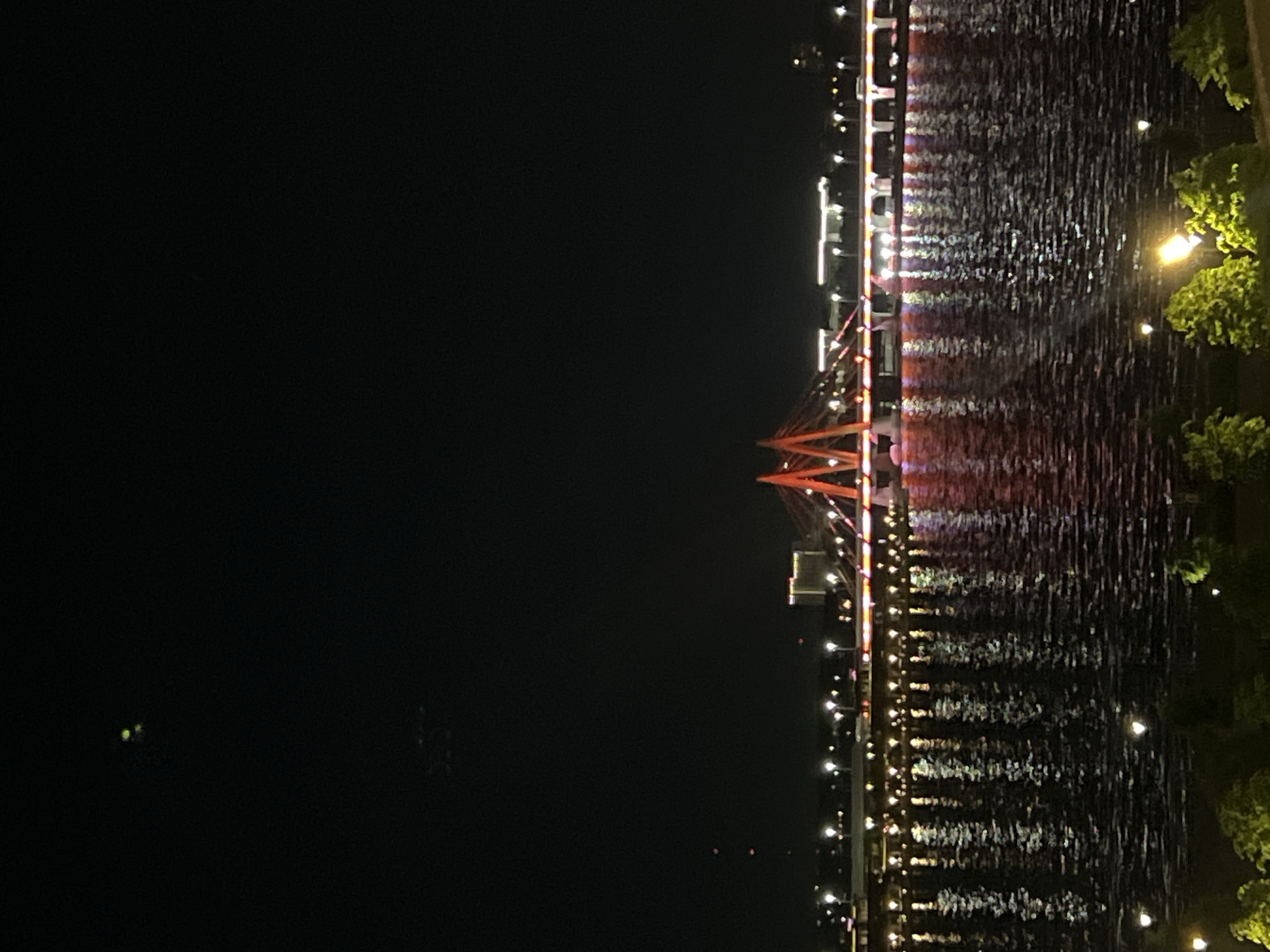 The image shows a nighttime view of the Milennium Bridge in Kazan, Russia. The bridge is illuminated with red and white lights. The lights are reflected in the water, creating a shimmering effect. There are also lights on the shoreline and on nearby buildings. Some trees are visible along the water's edge. The rest of the scene is shrouded in darkness.