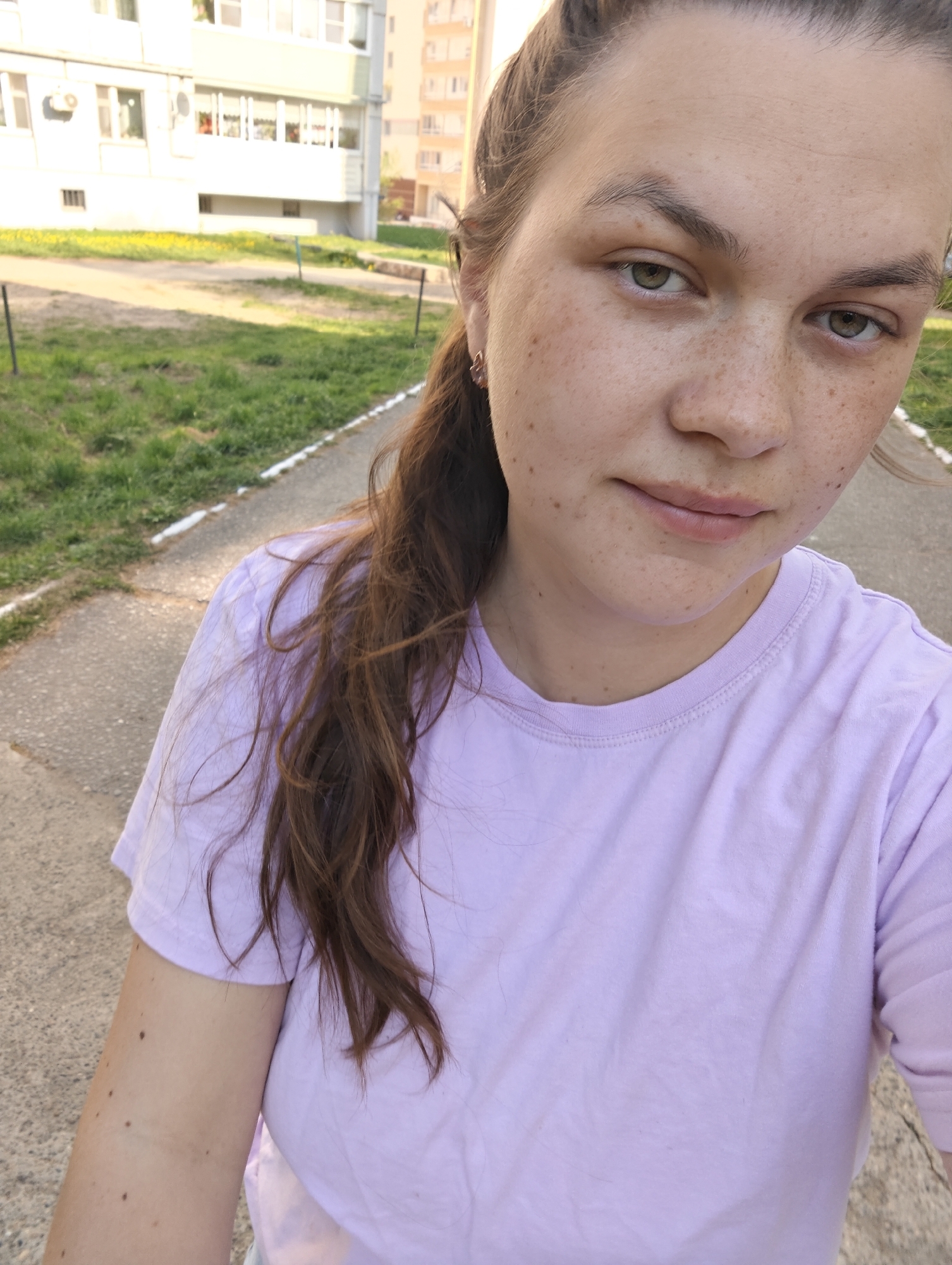 A woman is pictured in a selfie, outdoors on a paved walkway. A multi-story apartment building with white balconies and a green lawn are in the background. The woman is wearing a light purple t-shirt and has brown hair pulled back in braids with some strands loose. She has fair skin with freckles and is wearing small earrings. The setting appears to be in an urban area, and based on metadata, it's possible the location is Chaikovsky, Russia.
