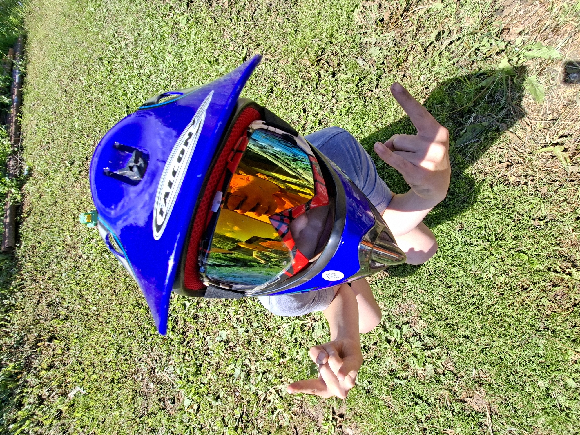 A person wearing a blue and silver motocross helmet with reflective goggles sits on a grassy area, raising both middle fingers in a gesture of defiance. The person's face is obscured by the helmet and goggles, but their arms and hands are visible. The background consists of green grass and a blurred wooden structure. The image is taken from a high angle, looking down on the person.