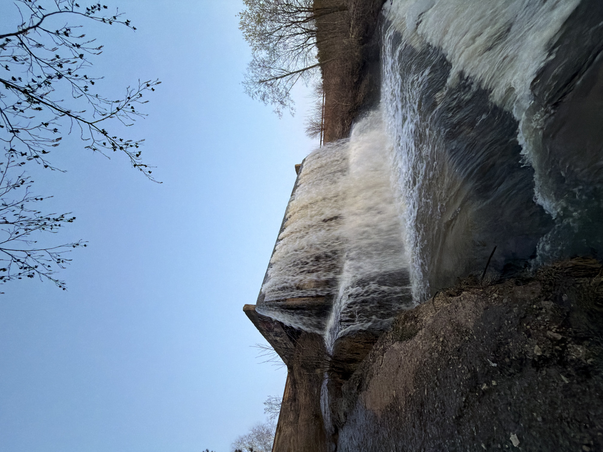 An image captures a wide waterfall or spillway, formed by water cascading over a substantial concrete dam or weir. The perspective is from below, looking upwards, giving the structure an imposing presence.
In the foreground, at the base of the waterfall, is a dark, rocky, and uneven ground or bank. The midground is dominated by the concrete structure, with large volumes of white, turbulent water flowing down its textured surface. The water spreads across a significant width of the dam, creating a powerful visual.
Above the dam, in the background, a treeline with bare or sparsely leaved trees is visible, suggesting a wooded area bordering the water body from which the water flows. The sky, occupying the upper left quadrant of the image, is a clear, light blue, indicating fair weather during the daytime. Bare branches and twigs from a tree extend into the frame from the far left, silhouetted against the sky.
No human activity or visible text is present. The scene depicts a blend of natural elements and engineered infrastructure, set in Kamennoye, Russia.