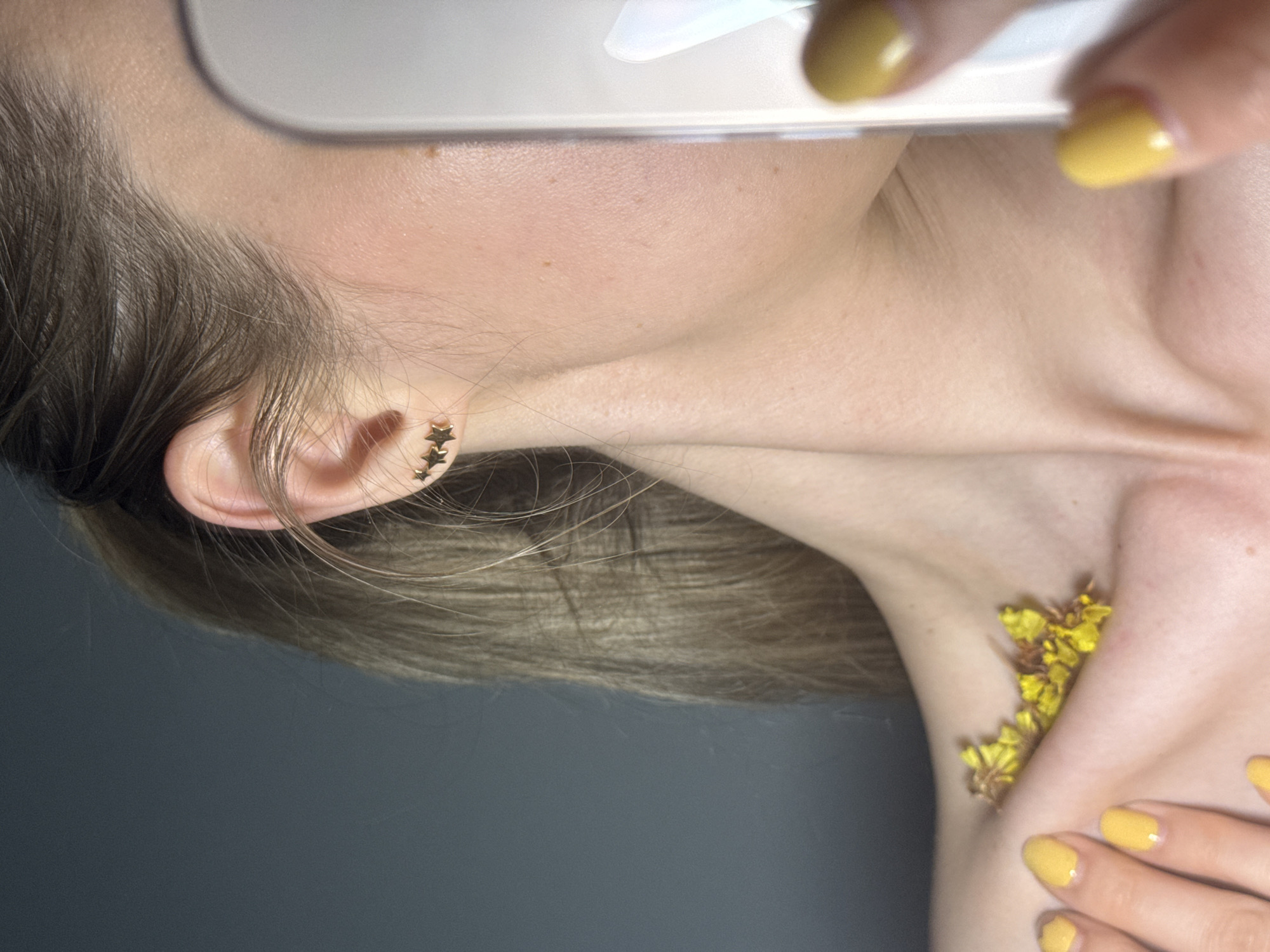 A close-up, artistically composed image features a person, likely female, positioned with her head to the left, showcasing her ear, neck, and upper chest against a plain, dark grey background. Her light brown hair is pulled back, revealing three small, golden, star-shaped earrings in her earlobe.
At the top of the frame, part of a white or light-colored smartphone or screen is visible, held by fingers painted with bright yellow nail polish. Another hand, also adorned with vibrant yellow nail polish, rests gently on the person's collarbone. Nestled into the hollow of the collarbone and neck are several small, dried yellow flowers, creating a subtle contrast with the fair skin.
The lighting is soft, highlighting the textures of the skin, hair, and the details of the accessories and flowers. The shot focuses on aesthetic elements, creating a delicate and curated impression. The overall activity appears to be a stylized portrait or a detail-oriented selfie. There are no discernible time of day, weather, or specific location cues within the image itself. No text is visible.