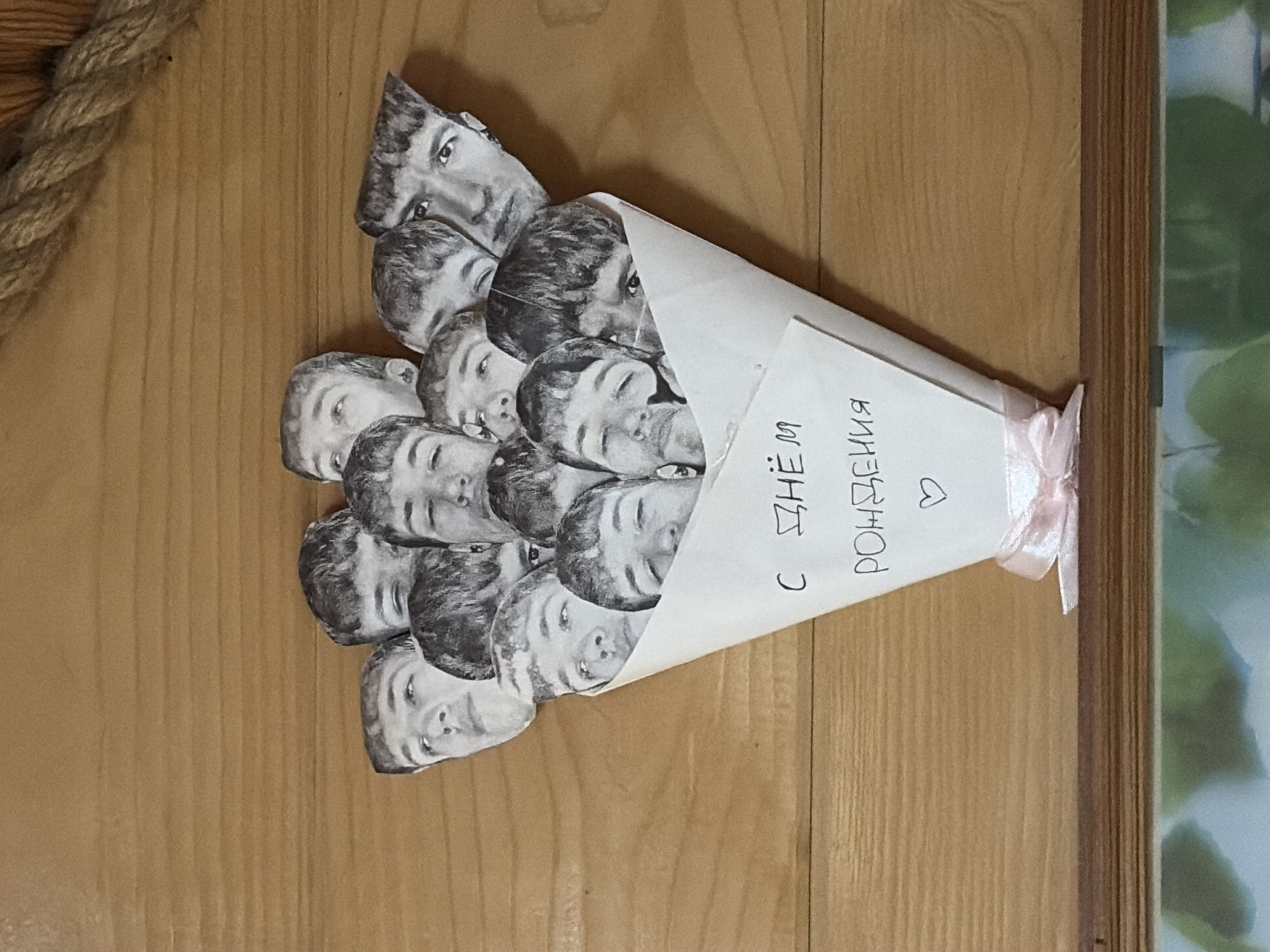 The image shows a bouquet of black and white photocopied faces arranged in a paper cone, tied with a pink ribbon. The text on the cone reads "С днём рождения" in Russian, followed by a heart symbol. The bouquet is placed on a light-colored wooden surface. A rope is visible in the upper left corner. The environment is indoors. There's no definitive information confirming the image was taken in Прикамский, Russia; this would need to be confirmed through other sources.