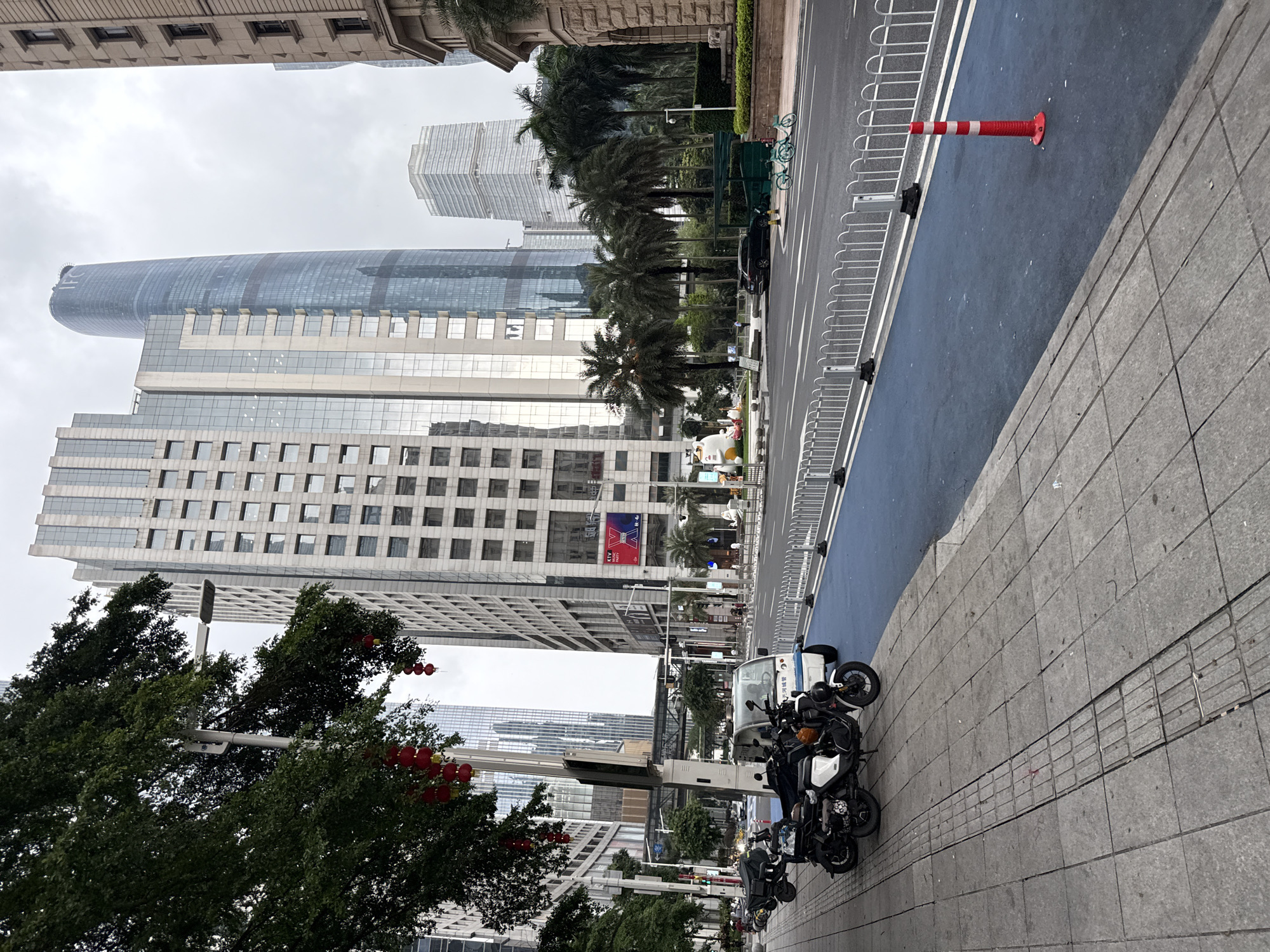 This image depicts a street scene in Tianhe District, China. The perspective is from a sidewalk looking towards a street and modern skyscrapers. Several motorcycles are parked on the sidewalk in the foreground. Beyond the motorcycles, a blue-colored lane, likely for cycling or a bus, runs alongside the road, separated by a white metal barrier. A red and white striped traffic cone is visible on the blue lane.
In the background, a series of tall, contemporary buildings with glass facades dominate the skyline. Some of these buildings have intricate architectural details, while others are more minimalist. Palm trees are interspersed among the buildings, suggesting a tropical or subtropical climate. The sky is overcast, indicating a cloudy or potentially rainy day.
The overall scene appears to be a typical urban environment during the day. There is no discernible human activity or any specific event taking place, beyond the presence of parked vehicles and the infrastructure of the street. Red lanterns are hanging from a tree in the left portion of the image, which might suggest a festive occasion or cultural decoration. Visible text on a digital billboard on one of the buildings reads "KTV Party". The weather appears to be gloomy, contributing to a neutral emotional tone.