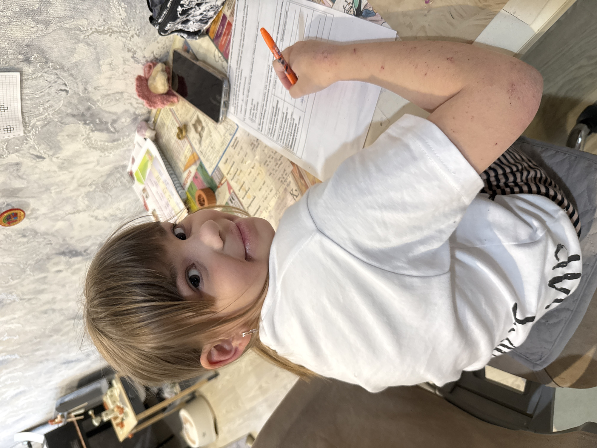 A girl with light brown hair sits at a table in Unknown, Unknown. She wears a white t-shirt and is positioned in a gray chair. She holds an orange pen and appears to be filling out a form. Various items such as a phone, a crocheted item, papers, and other unidentifiable objects are scattered across the table. The background includes a marbled surface and glimpses of other furniture.