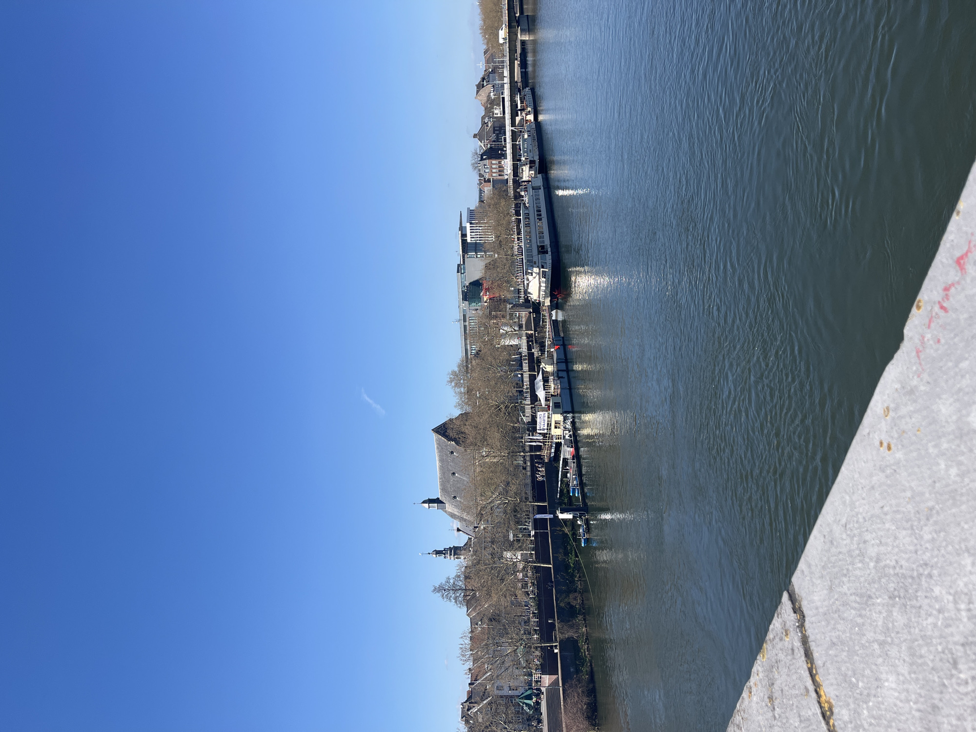 The image depicts a view of the Maas river in Maastricht, Netherlands, looking towards the city center. Several boats are docked along the riverbank. Buildings, including a church with a steeple and a sloped roof, line the waterfront. Bare trees suggest it is late autumn or early spring. The sky is clear and blue. The photograph appears to be taken from a bridge, with part of a concrete railing visible in the bottom right corner.