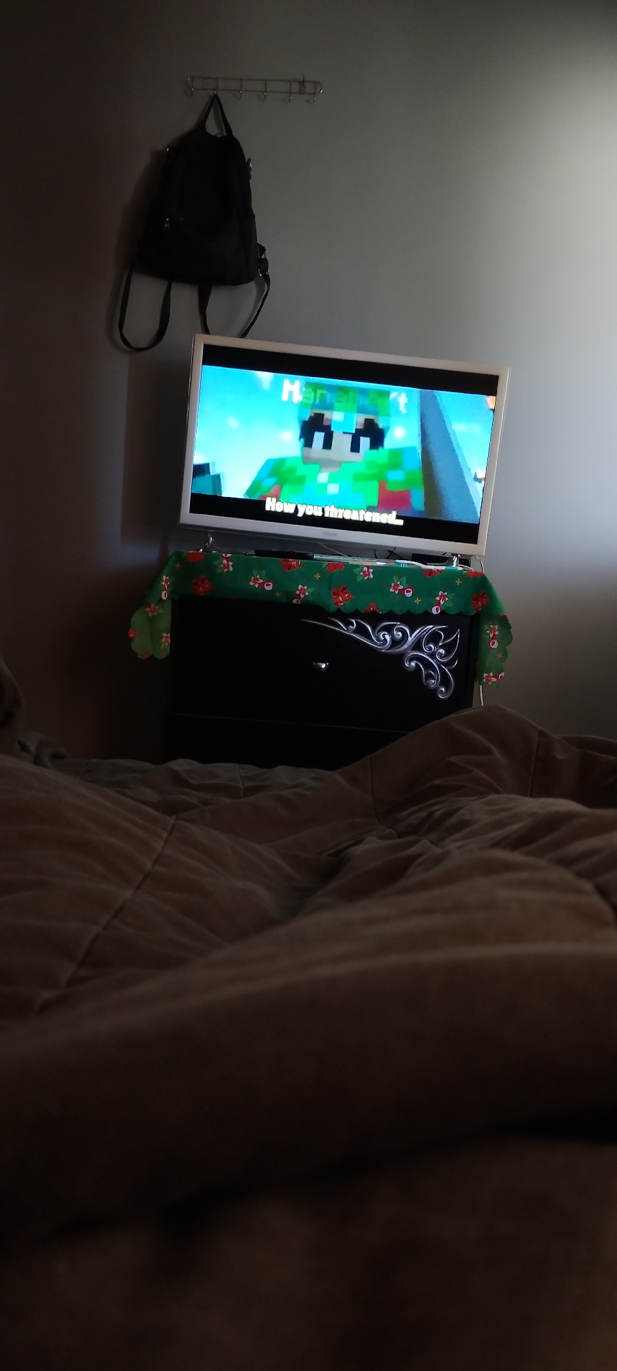 In a dimly lit room, likely a bedroom, a flat-screen television is mounted on the wall above a black dresser. The television displays a video game character with a green shirt and blue hair, and text that reads "How you threatened...". A patterned cloth with red and white flowers is draped over the dresser. A black backpack hangs on a hook on the wall to the left of the television. The foreground is dominated by a close-up of a rumpled, brown comforter, suggesting the viewer is in bed. The scene takes place in an interior setting, possibly a home in Zimnitsa, Bulgaria, given the context provided.