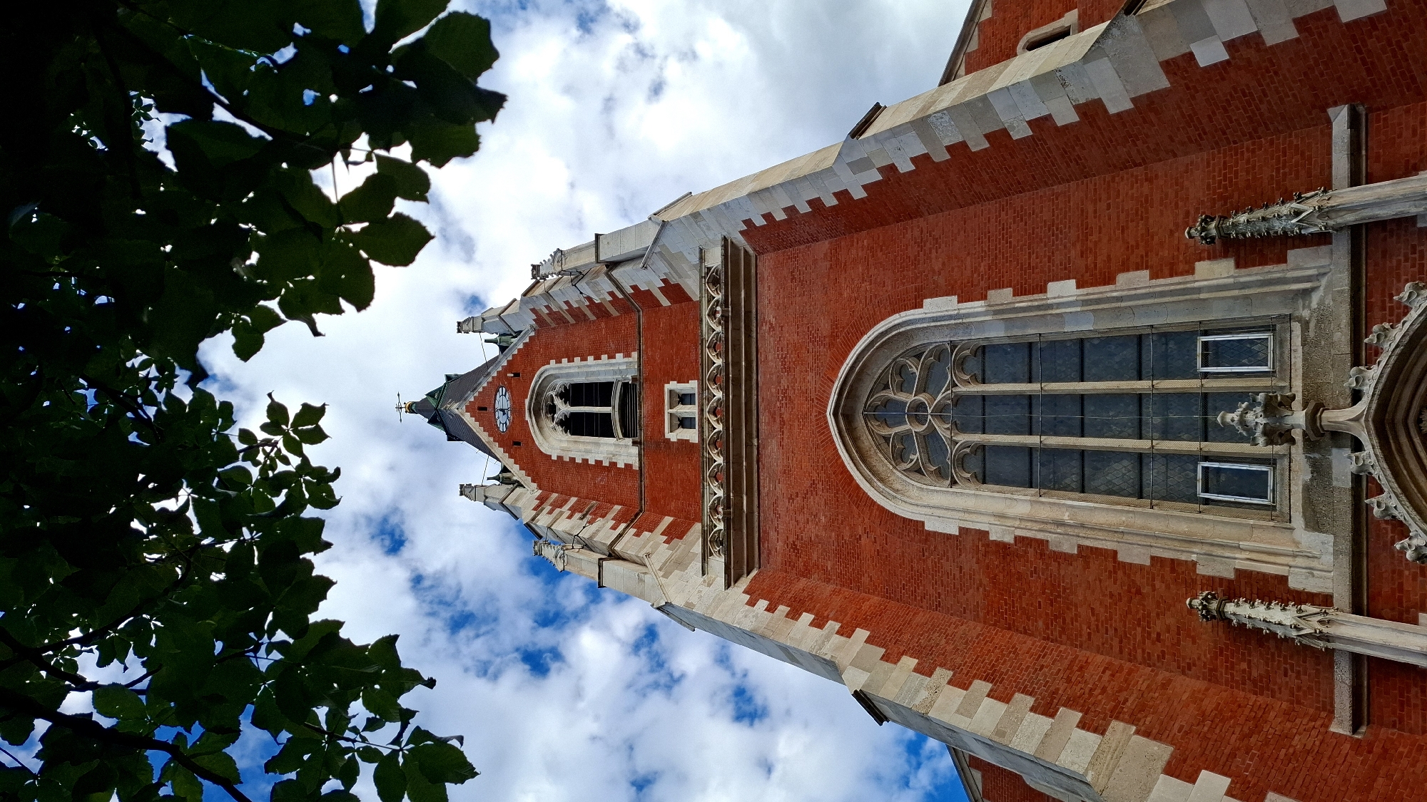 The image shows a low-angle view of the brick tower of St. Stephen's Cathedral in Vienna, Austria, framed by dark green tree branches and a cloudy blue sky. The tower is constructed of red brick with white stone accents. Gothic-style arched windows with decorative tracery are visible, as are smaller rectangular windows. The top of the tower features a steep roof and a spire with a cross. A clock face is visible on one side of the tower near the top. The overall impression is one of architectural grandeur and historical significance against a natural backdrop.