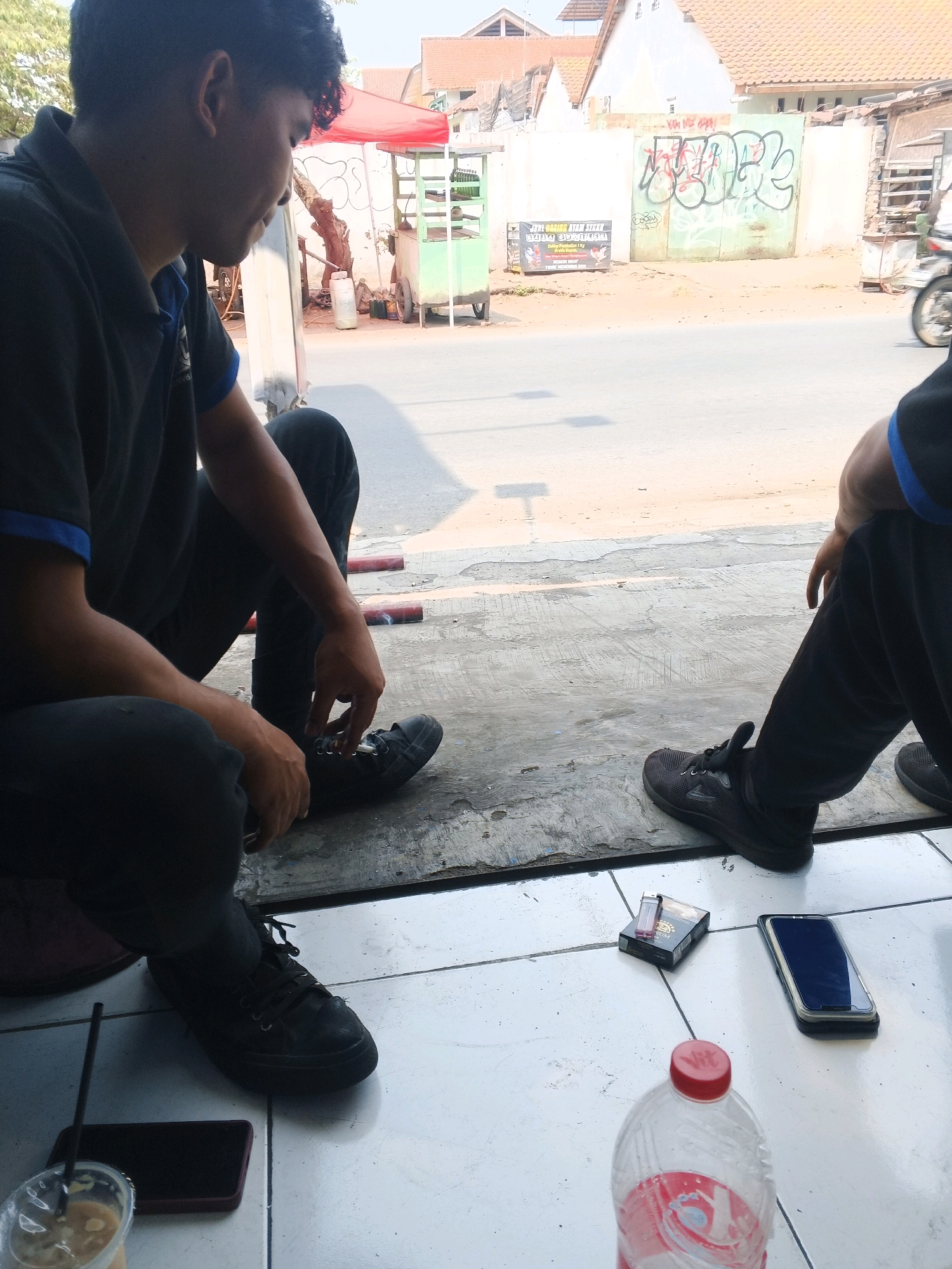 The image shows two men sitting on a tiled floor in front of a street in Taman, Indonesia. The man on the left is wearing a black polo shirt and black pants. He is looking down at his shoes, one of which he is tying. The man on the right is wearing a black shirt and black pants. He is leaning back and looking away from the camera.
On the floor in front of the men are various objects, including a pack of cigarettes, a lighter, a smartphone, a plastic bottle of water, and a cup of iced coffee with a straw. In the background, there is a street with a motorcycle passing by. Beyond the street, there are buildings with tiled roofs and graffiti on a wall. There is also a red tent and a green food cart. The lighting suggests it is daytime.