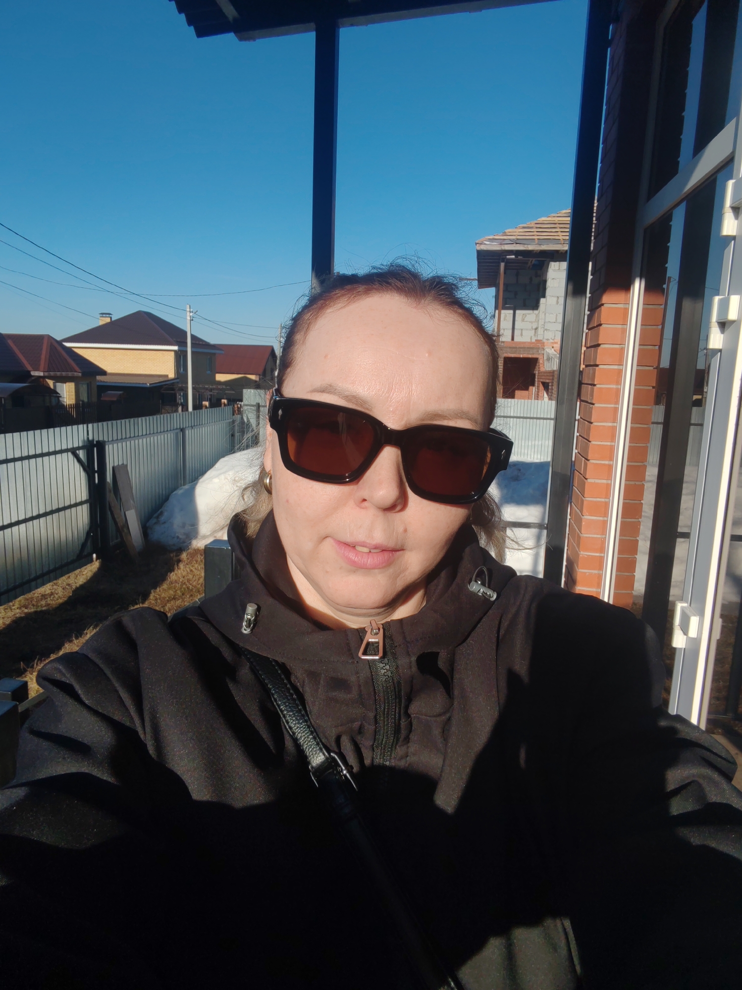 The image is a selfie of a woman wearing dark sunglasses and a black jacket with a zipper. The background shows a residential area with houses, a fence, and a building under construction with a roof. The sky is clear and blue. There is a partial structure in the foreground. The location is indicated as Chaikovsky, Russia.