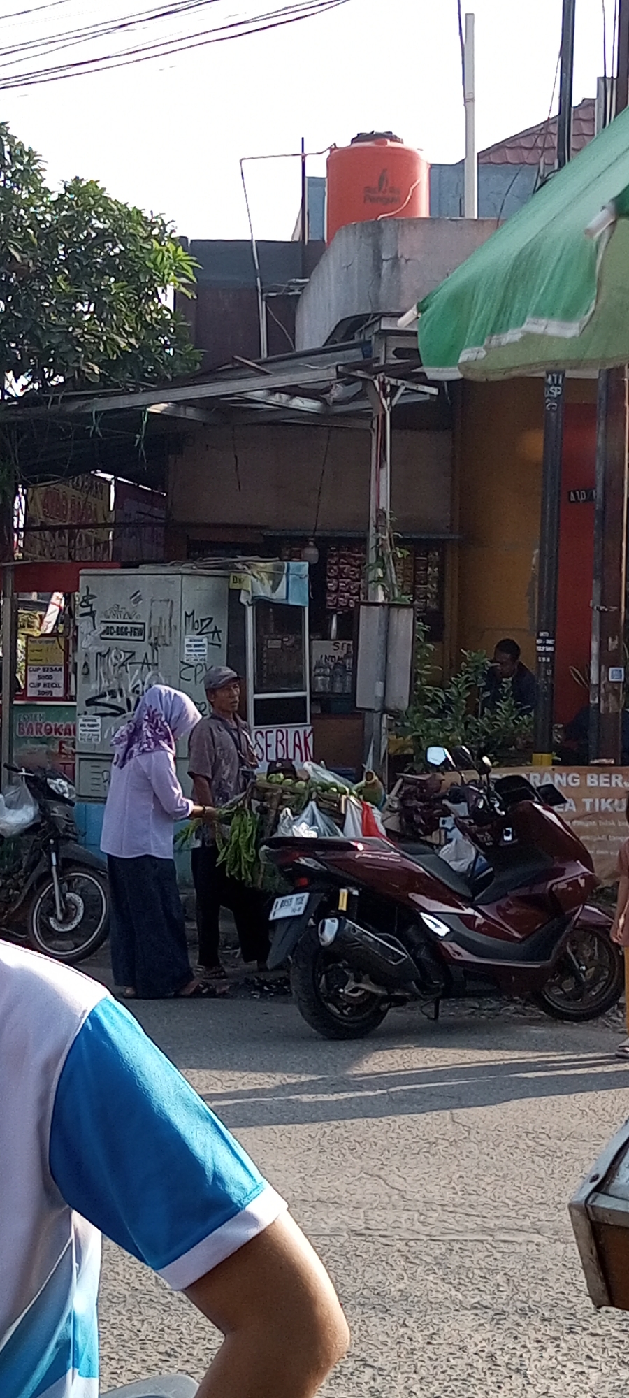 In the city of Setu, Indonesia, a street scene unfolds. The foreground is dominated by the arm of a person wearing a blue and white shirt, partially obscuring the view of the street. Behind them, a dark red motorcycle is parked, its license plate reading "B 8533 YCE". To the left, a dark-colored motorcycle is also visible.
The mid-ground features two individuals interacting. A woman wearing a light purple hijab and a long skirt stands facing a man. The man, wearing a grey cap and a dark shirt, appears to be holding a bundle of green vegetables. They are positioned in front of a small food stall or shop. The stall has a metal frame and is adorned with various items and signage, including what appears to be a refrigerator with graffiti. Another person can be seen sitting behind the counter inside the stall.
In the background, a large orange water tank sits atop a concrete structure. The environment suggests a bustling urban setting with buildings and overhead wires. The lighting indicates it is daytime, with shadows cast on the asphalt road.