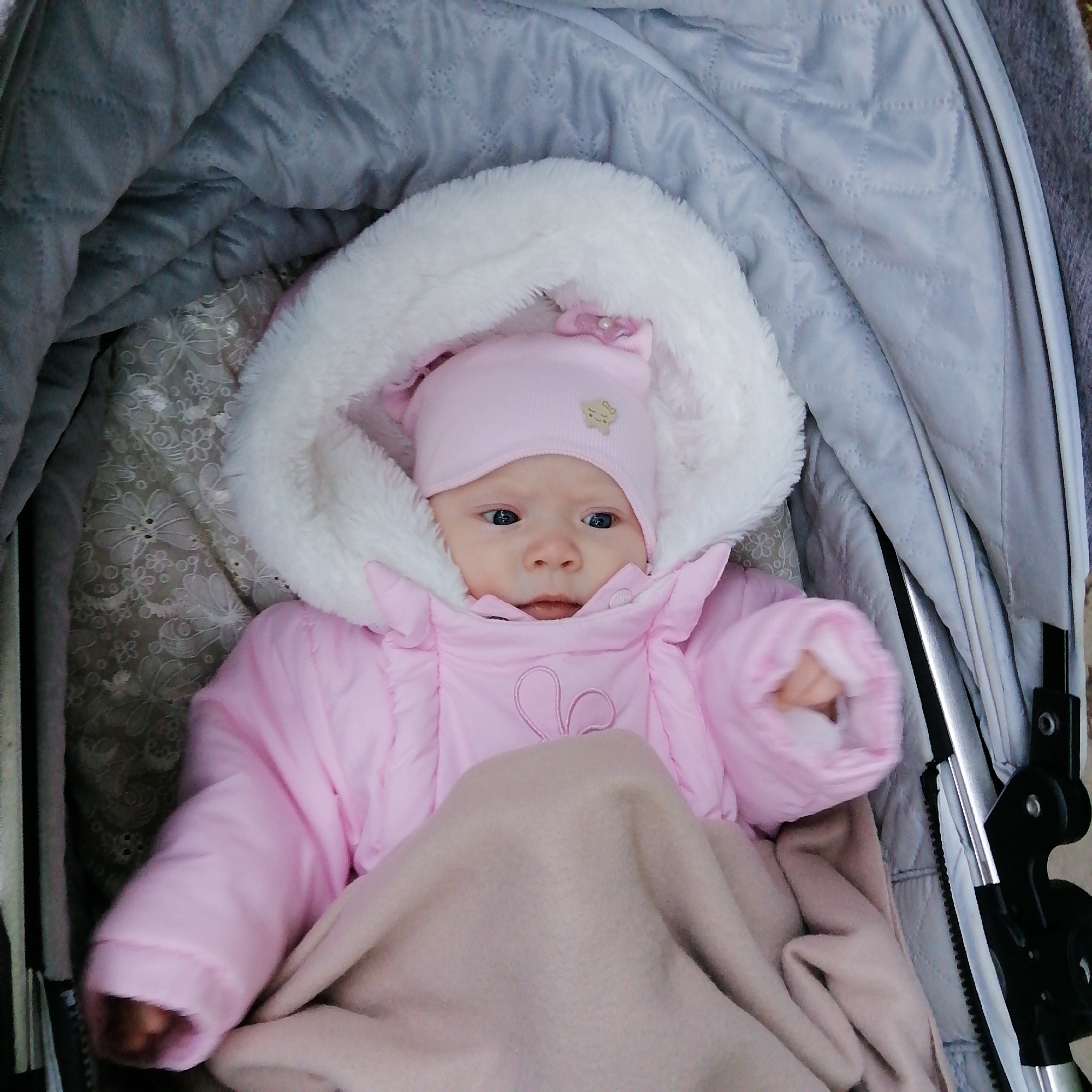 A baby wearing a pink snowsuit with a white fur-trimmed hood and a matching pink hat is lying in a stroller. The stroller is lined with a grey quilted fabric and a patterned fabric, and the baby is partially covered by a beige blanket. The stroller is in an unknown location in Трубачеевка, Russia.