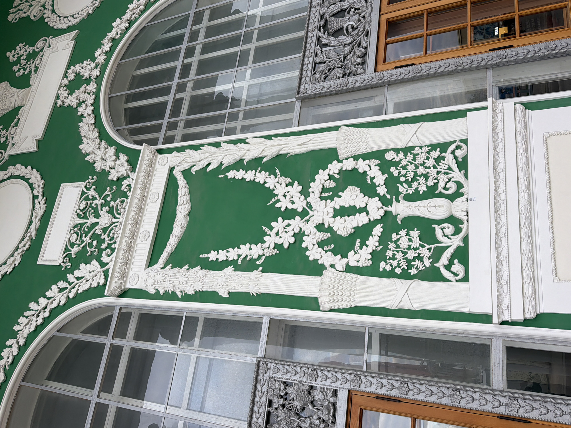 The image shows a fragment of architectural decor, likely part of a building facade. The background is painted a deep green, and overlaid with ornate white plaster or stucco relief. The relief includes floral and foliate patterns, geometric motifs, and decorative borders.
Two large, arched windows are visible, each divided into multiple panes by a grid of mullions. One window has a decorative metal frame with what appears to be a wooden-framed window set within. The scene implies an exterior view of the architecture.
The architecture style suggests a historical building located in a city. Given the architectural style, the location is possibly Moscow, Russia, based on the presence of similar architectural ornamentation in that city. However, without additional context or information, it is impossible to confirm with certainty.