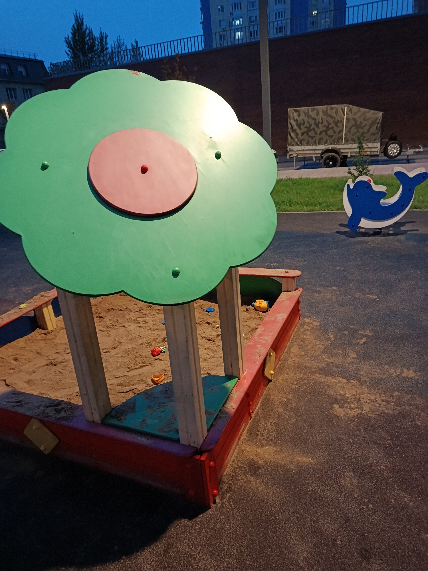 The photograph, likely taken at dusk, shows a children's playground. The primary focus is a sandpit with a green, cloud-shaped shade structure. The sandpit's border is red with yellow accents. Inside, the sand contains a few scattered toys.
To the right of the sandpit is a blue dolphin-shaped rocking toy. In the background, a brick wall is visible, topped with a metal railing. Beyond the wall, there is grass, a trailer covered in camouflage fabric, and a multistory apartment building.
The location is likely in Moscow, Russia, given the style of the buildings. No people are visible in the scene.