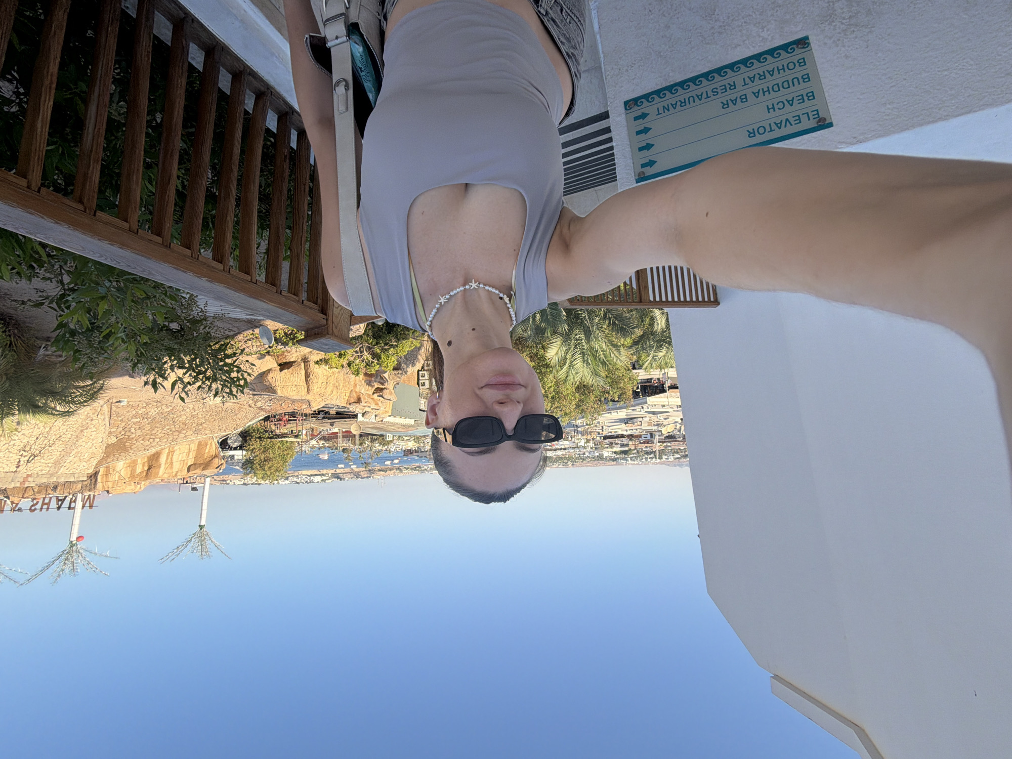 A young woman with dark hair pulled back and wearing sunglasses takes a selfie outdoors. She is wearing a light gray tank top, a pearl necklace, and jeans. Behind her, a wooden railing separates her from a view of a coastal city with buildings and boats on the water. Palm trees and rocky structures are also visible. A sign on a white wall in the foreground indicates directions to an elevator, beach, Buddha Bar, and Boharat Restaurant. The setting is Sharm El Sheikh, Egypt.