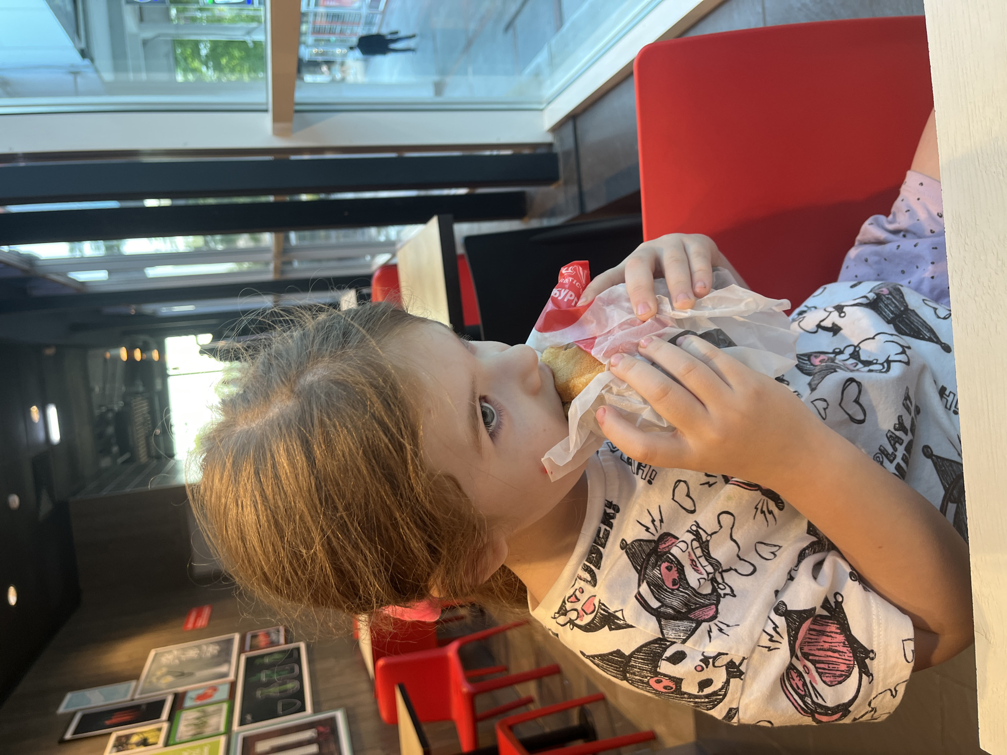 A young girl is eating a small sandwich wrapped in white paper in a brightly lit restaurant. She is sitting at a table with a white surface and a red chair is visible to her right. The restaurant appears to have a modern design with a large window showing a street scene outside. A few framed pictures are hanging on a dark wall to the left. The girl has brown hair pulled back with a pink scrunchie and is wearing a white t-shirt with a cartoon print. The overall impression is that of a casual meal in a public space, likely a fast-food establishment.
