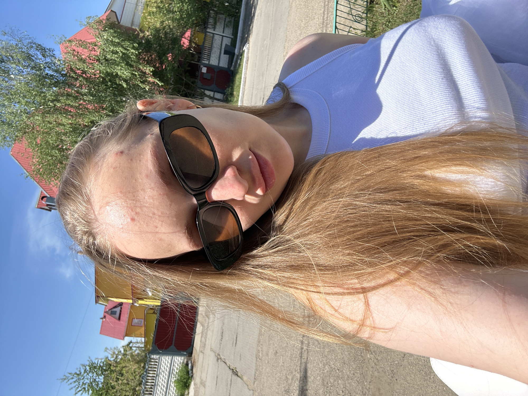 A photograph shows a fair-skinned person with long, light brown hair, wearing dark sunglasses and a white tank top. The person's face is angled slightly, and they are in an outdoor setting. In the background, there is a road, a fence, trees, and buildings. One building has a red roof, another is yellow with red accents, and there's a glimpse of a third building with graffiti. The photo appears to be taken in the city of Chaikovsky, Russia.