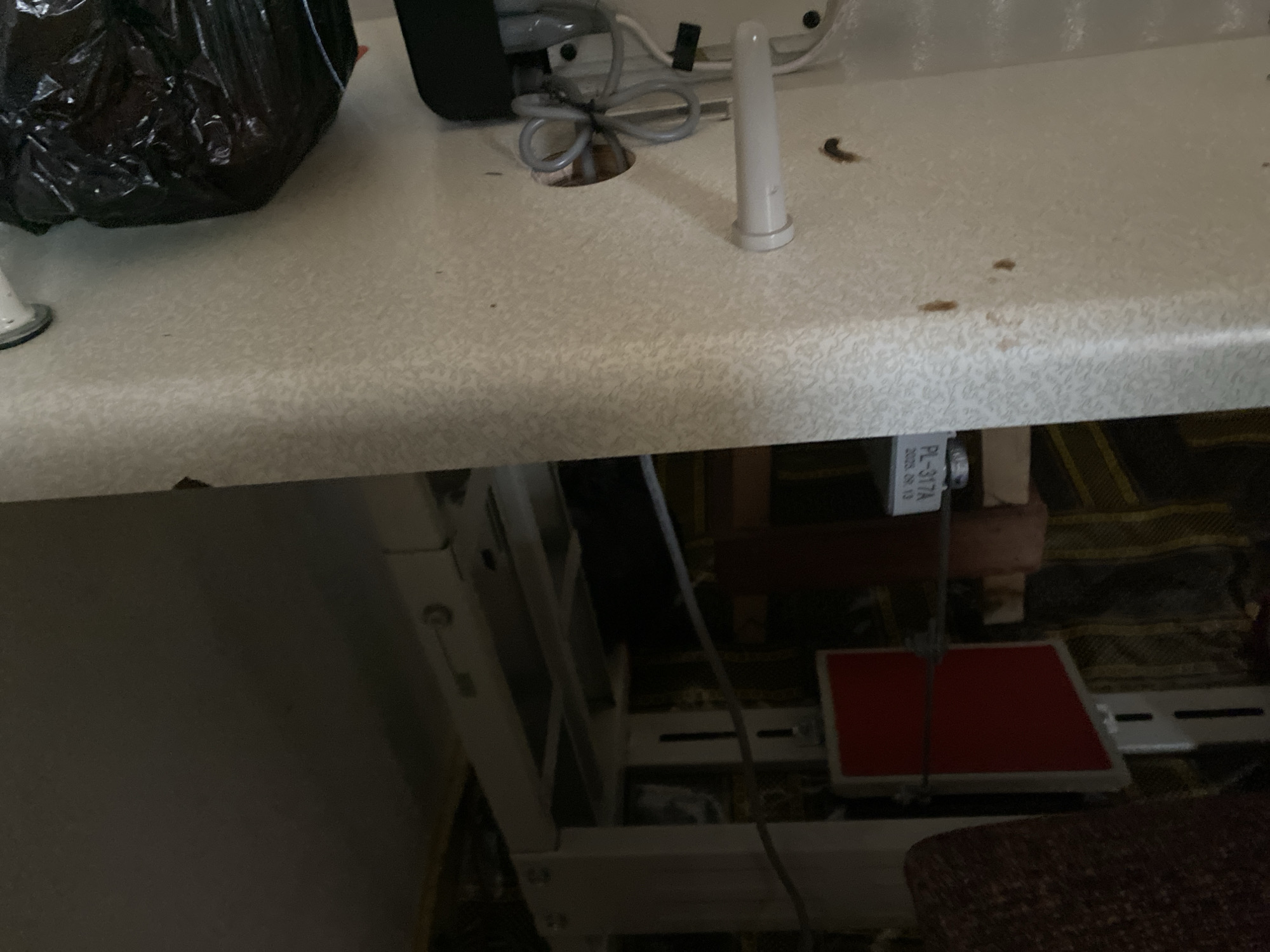 The image shows a close-up, overhead view of a white sewing machine table. On the left side of the table, a large black plastic bag is visible, obscuring a portion of the surface. To the right of the bag, a white, cylindrical object, likely a spool pin or a part of the sewing machine, stands upright. A hole is present in the table, through which a coiled gray cord emerges. There are several small brown specks on the table's surface, possibly dirt or debris.
The bottom half of the image reveals the underside of the sewing machine table and the structure supporting it. A white metal frame forms the base, with various components and cables visible. A red square object, possibly a presser foot or a component of the sewing machine, is situated on the frame.
A small white label is attached to a metal component of the sewing machine, bearing the text "PL-317A" and what appears to be a date: "2018.09.13". The lighting in the image suggests an indoor setting, likely during daylight hours. No people are visible in the frame. The overall scene depicts a workspace related to sewing. The provided location context of Jauro Yinu, Nigeria, indicates the general geographical setting.