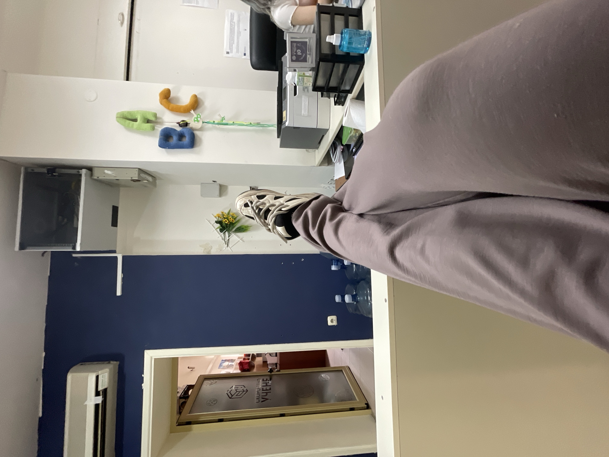 The image captures an indoor scene from a low, first-person perspective, looking across a counter or desk. In the immediate foreground, a person's legs, clad in light mauve sweatpants, are propped up on a light beige counter surface. The right foot, wearing a beige and black athletic shoe with white laces, rests on the counter.
Beyond the counter, the room features a dark blue wall on the left and a white wall on the right. High on the dark blue wall, a white split-system air conditioning unit is mounted. Below it, near the floor, an open doorway with a light-colored frame reveals an adjacent room. The door is frosted glass, featuring a geometric logo and the Cyrillic text "СКОРОСТНО УЧЕНЕ" (Speed Learning). Through the doorway, red objects and shelves are partially visible. On the floor near the dark blue wall, several large, clear plastic water bottles are present. A white electrical outlet is visible on the dark blue wall.
On the white wall to the right, three plush letters – a green 'A', a blue 'B', and an orange 'C' – are attached, with a decorative green string hanging below them. A small cluster of artificial yellow flowers is positioned on a surface near these letters. Below these decorations, a white desk holds a grey and white printer, a small transparent frame displaying "УЧЕНЕ 10" (Learning 10) in Cyrillic, a black multi-compartment desk organizer, and a blue spray bottle. A person with light-colored hair and wearing a white top is partially visible seated behind this desk on the far right of the frame. The ceiling is white, and a white rectangular fixture is visible at the junction of the white wall and ceiling on the right side. The overall environment suggests an office or learning institution in the city of Unknown, Unknown.