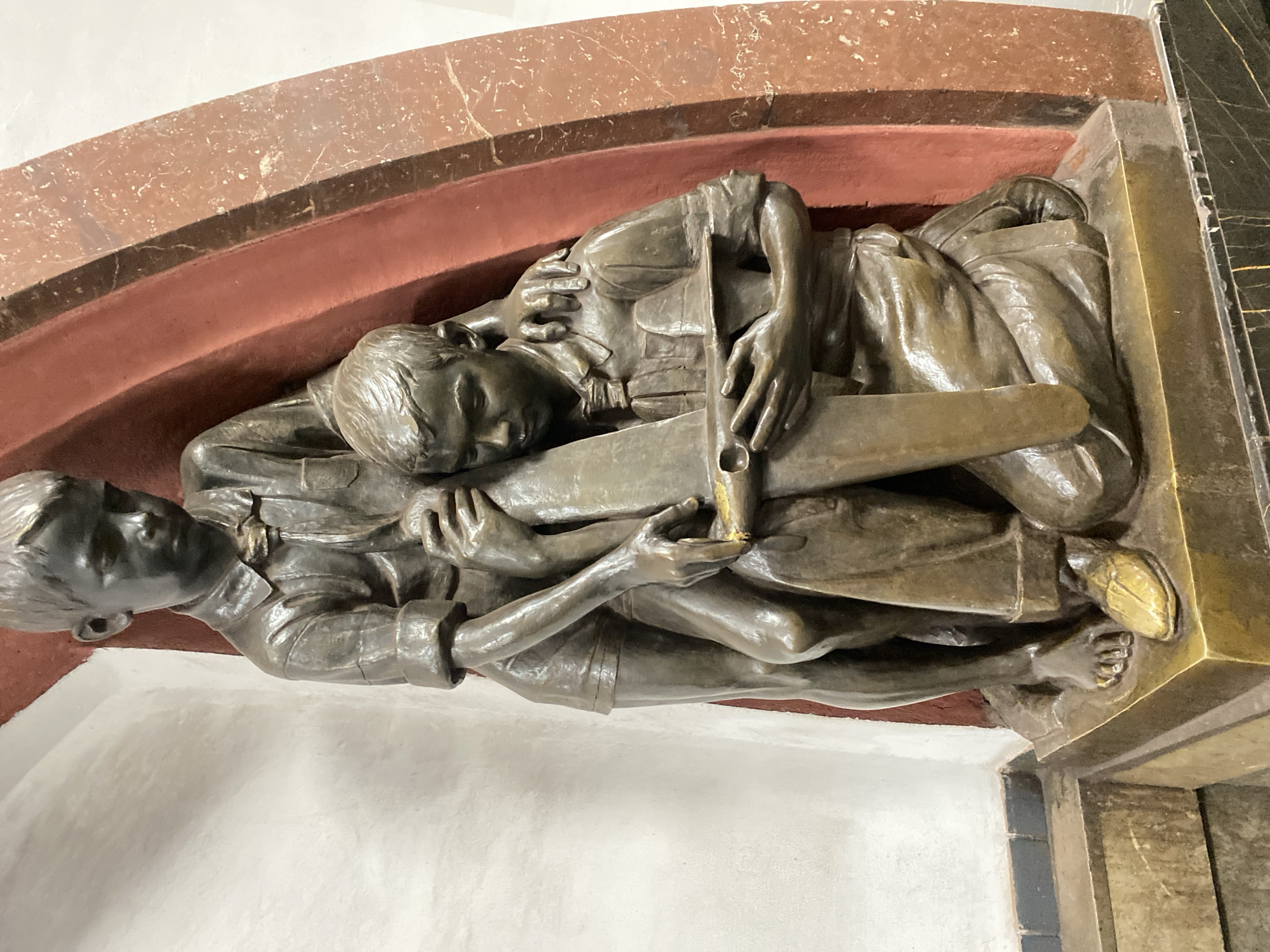 The image depicts a bronze sculpture of two male figures lying together. The upper figure rests his head on the shoulder of the lower figure, clutching a wooden airplane propeller. Both are dressed in what appears to be early 20th-century attire, possibly aviation uniforms. The sculpture is mounted on a wall within an arched niche, trimmed with red and brown stone. Adjacent is a section of dark, patterned stone. The location is identified as Moscow, Russia.