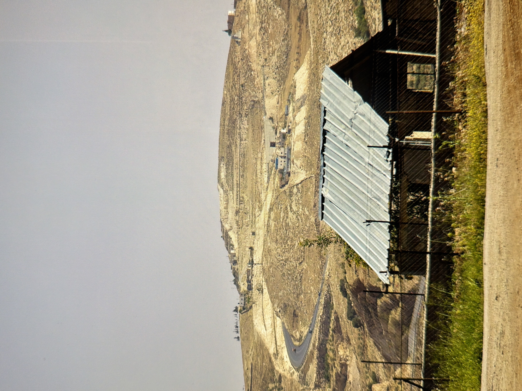 The image depicts a rural landscape with a winding road leading up a hillside. In the foreground, a small, dilapidated structure with a corrugated metal roof is visible, enclosed by a chain-link fence. Beyond this structure, the dry, rocky terrain of the hillside is dotted with scattered buildings and vegetation. The sky is clear and pale. The scene is a generic rural setting and does not provide any information that would indicate it is located in Kyiv, Ukraine.
