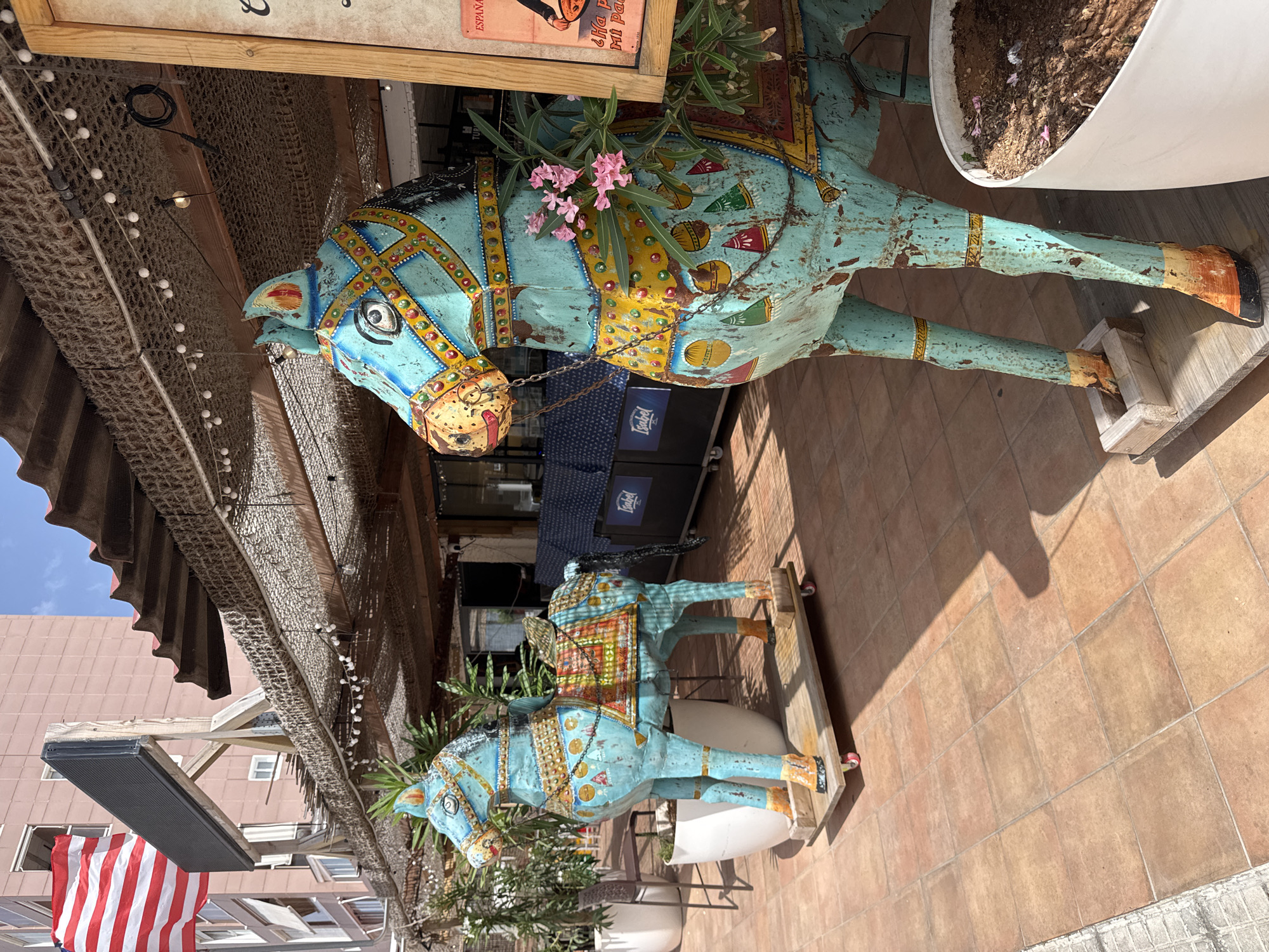 The image depicts an outdoor seating area with two decorative horse statues in the foreground. The statues are light blue with intricate yellow, red, and green patterns. They are weathered, showing signs of rust and peeling paint. The statue on the right has pink flowers growing from its saddle area. In the background, there is a building with a pink facade and an American flag flying. A corrugated roof covers the seating area, adorned with string lights. Tables and chairs are visible in the distance, along with a blue banner displaying the word "Isabel." The ground is paved with terracotta tiles. The location is identified as being in Gandia, Spain, likely an establishment with outdoor seating, possibly a restaurant or bar. There are no people visible in the image. The scene is brightly lit by sunlight, casting shadows on the tiled ground.