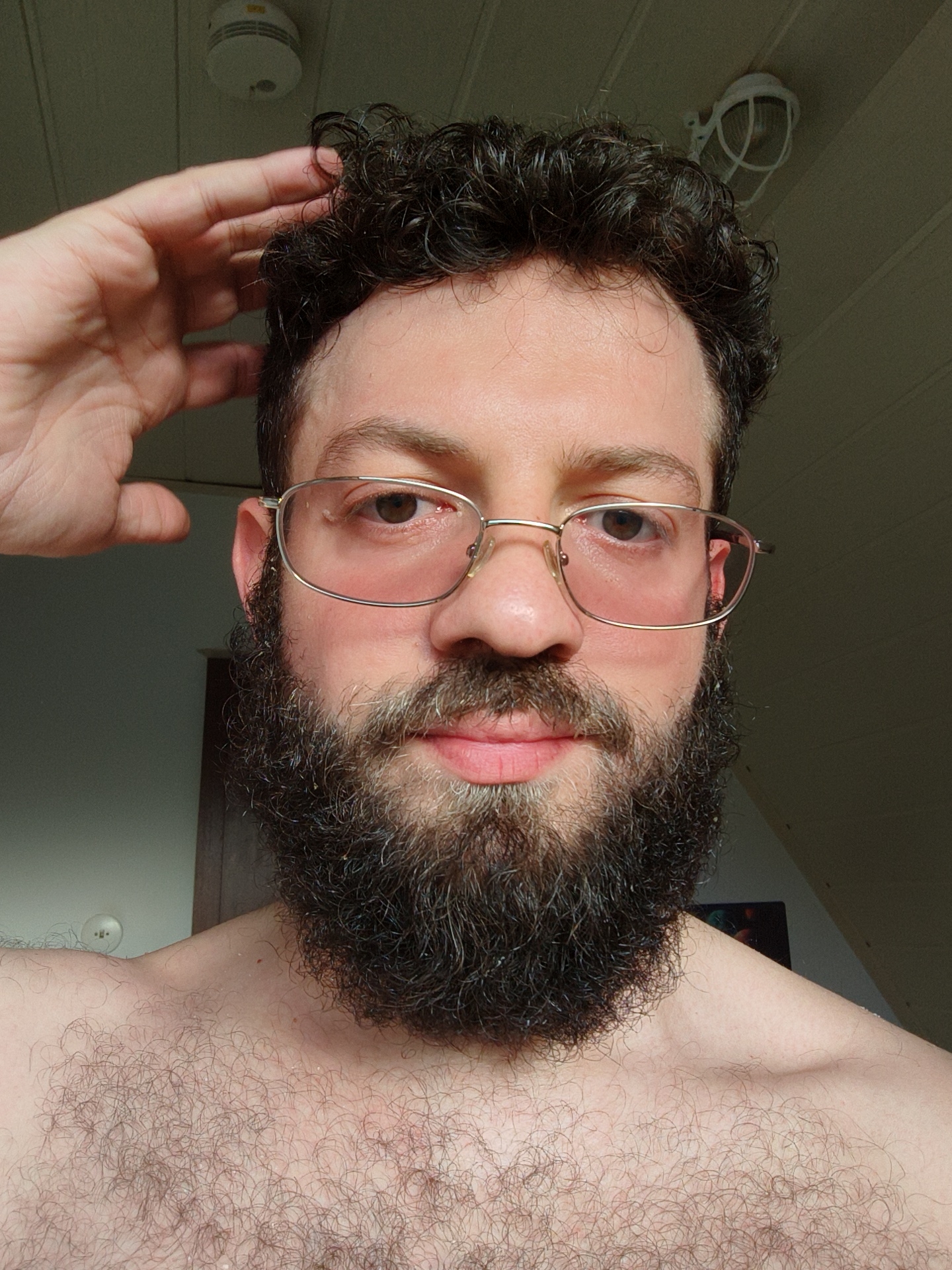 A medium close-up shot depicts a fair-skinned male with a dark, full beard and curly dark hair. He is wearing gold-rimmed glasses with a slight rose tint. His right hand is raised, touching his hair near his temple. He appears shirtless. The background consists of a plain white ceiling with a smoke detector and an exposed light fixture with a wire cage. Parts of a wall and door are visible behind him. The location is not discernible from the image. There is no evidence in this image to suggest that the picture was taken in Diepholz, Germany.