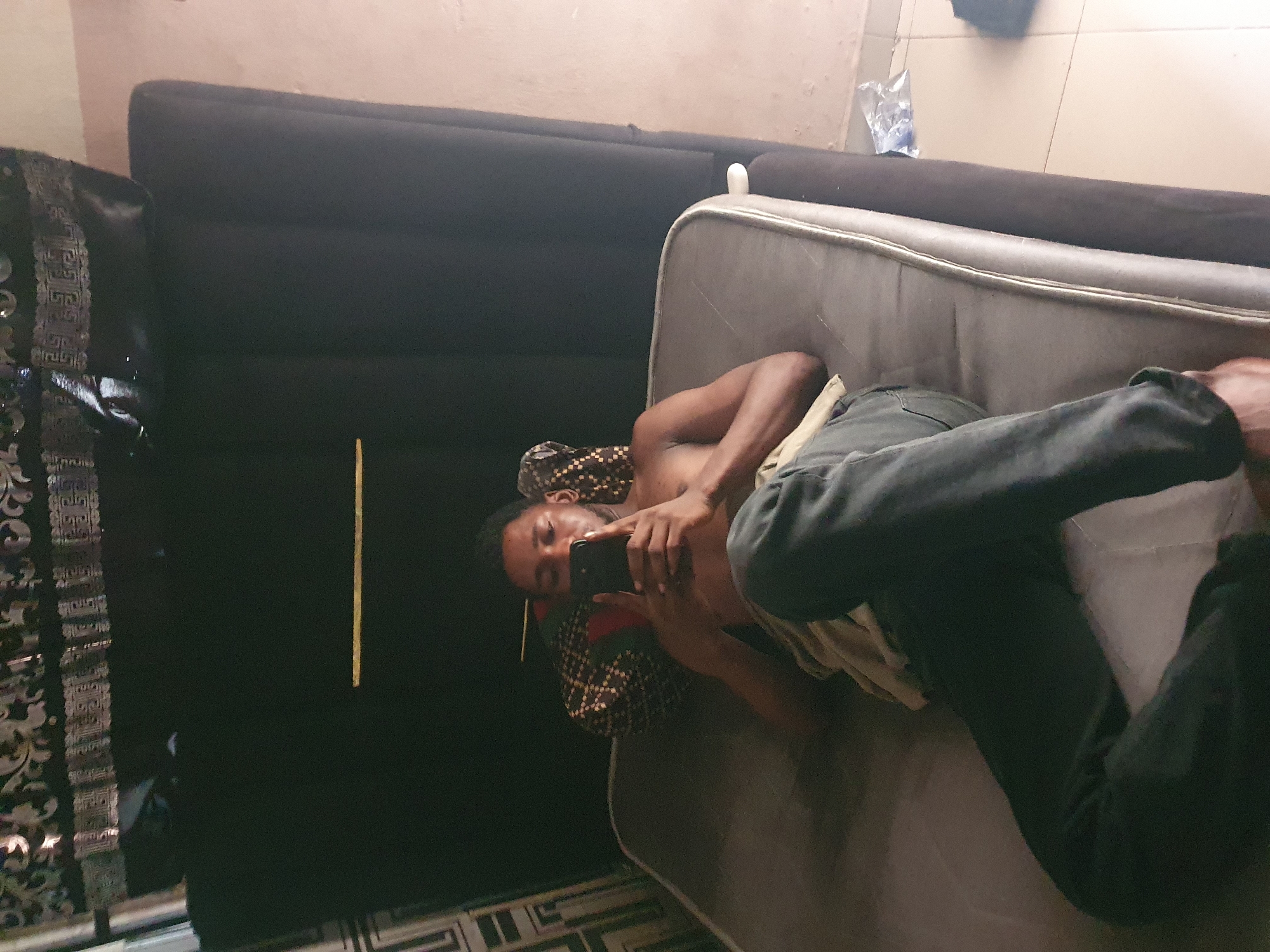 A shirtless young Black male is lying on a mattress, propped up by a pillow. He is holding a smartphone in his hands and looking at it. He is wearing dark gray pants. The mattress is on top of a dark gray headboard or frame. The background is a dark wall with some patterned fabric hanging from the top. The floor is tiled with beige tiles. There is a crumpled plastic bag on the floor to the right of the mattress. The overall lighting is dim.