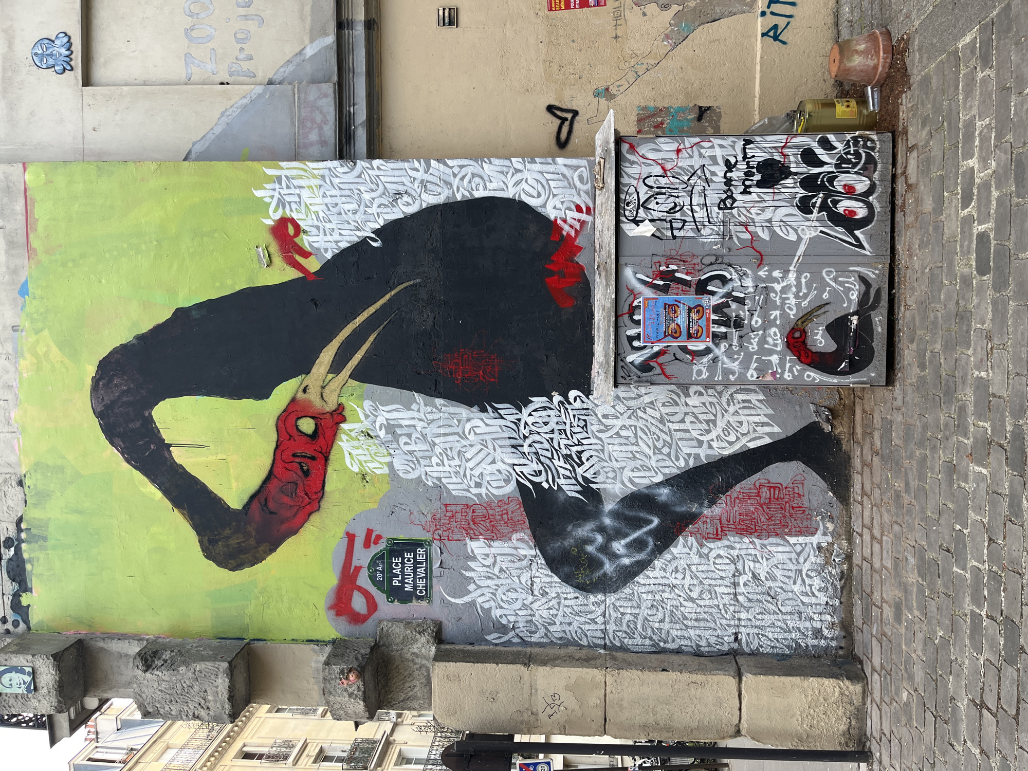 The image captures a vibrant scene of street art in Paris, France, specifically at Place Maurice Chevalier in the 20th arrondissement. Dominating the left portion of the frame is a large mural featuring a stylized bird-like figure with black body and a red head and neck. The background is a mix of lime green and gray. The mural is complemented by white, swirling calligraphic text and other red markings that appear around the bird.
To the right of the mural, a utility box or door is covered in additional graffiti, including a smaller version of the same bird figure, overlapping tags and scribbles, and a colorful flyer. At the top of the utility box sits a broken clay pot and a partially visible clear plastic bottle.
The artwork and utility box are flanked by stone architectural elements, indicating the site is likely on or near a building's facade. The ground is paved with interlocking cobblestones, suggesting a pedestrian zone or alley. Fragments of other buildings and a street lamp can be seen in the lower portion of the frame. The overall environment is urban and shows a high concentration of street art.