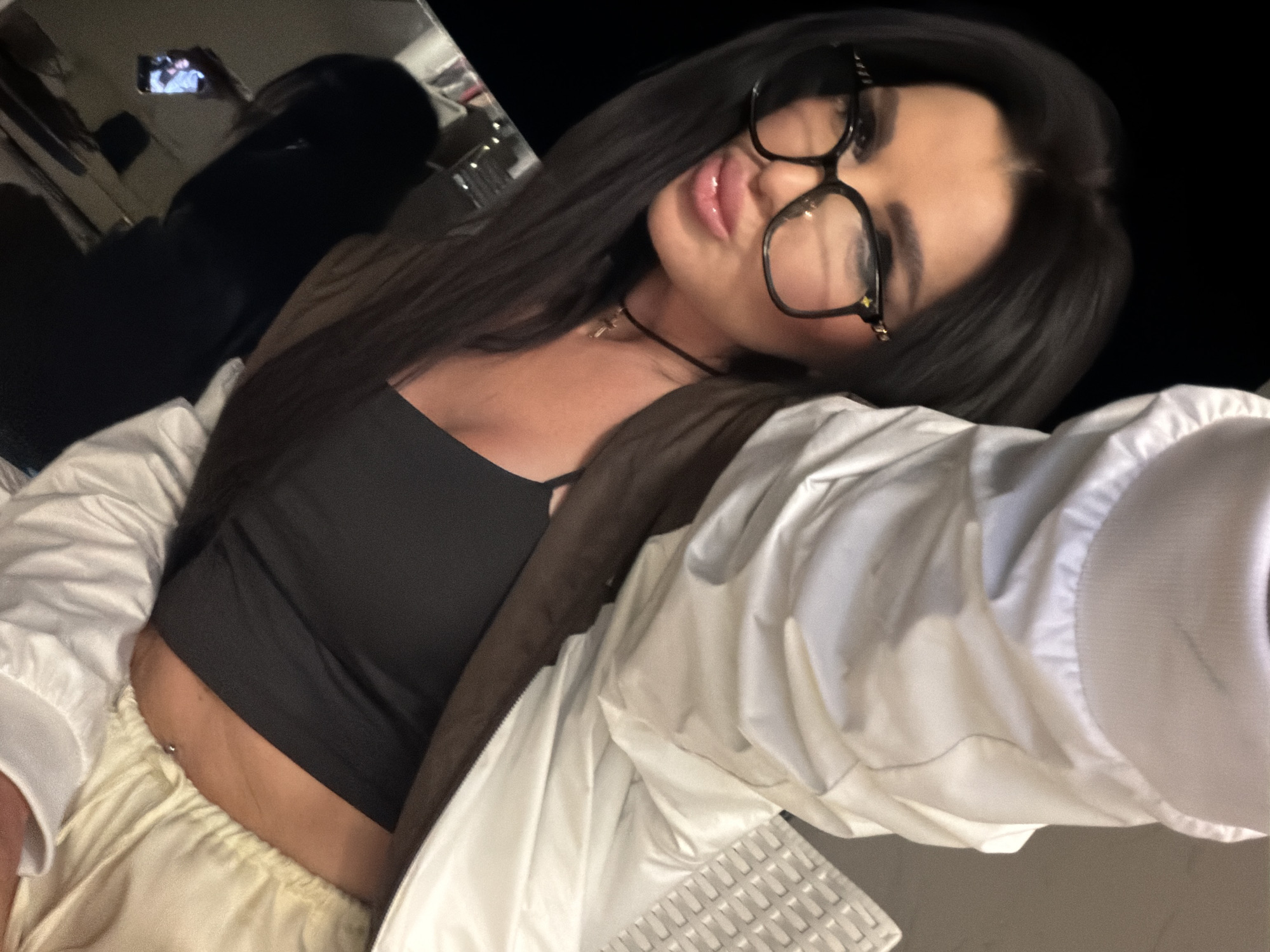 An angled, close-up selfie shows a young woman with long, dark hair, looking towards the camera with pursed lips. She wears large, dark-framed glasses with a visible gold "LV" logo on the temple piece.
She is dressed in a black crop top, revealing a belly button piercing. A black choker necklace adorned with a small gold cross pendant is visible around her neck. Over the crop top, she wears a light-colored, possibly off-white or cream, zip-up jacket or windbreaker. Her lower attire appears to be light-colored, matching pants or shorts.
The background is dark and out of focus, suggesting an indoor setting or a dimly lit environment, likely at night. In the upper left corner, a reflection in what appears to be a mirror shows a phone screen displaying an image, possibly indicating how the selfie was taken or the presence of another device.
The overall impression is casual and confident. The location context provided is Boreti, Montenegro.