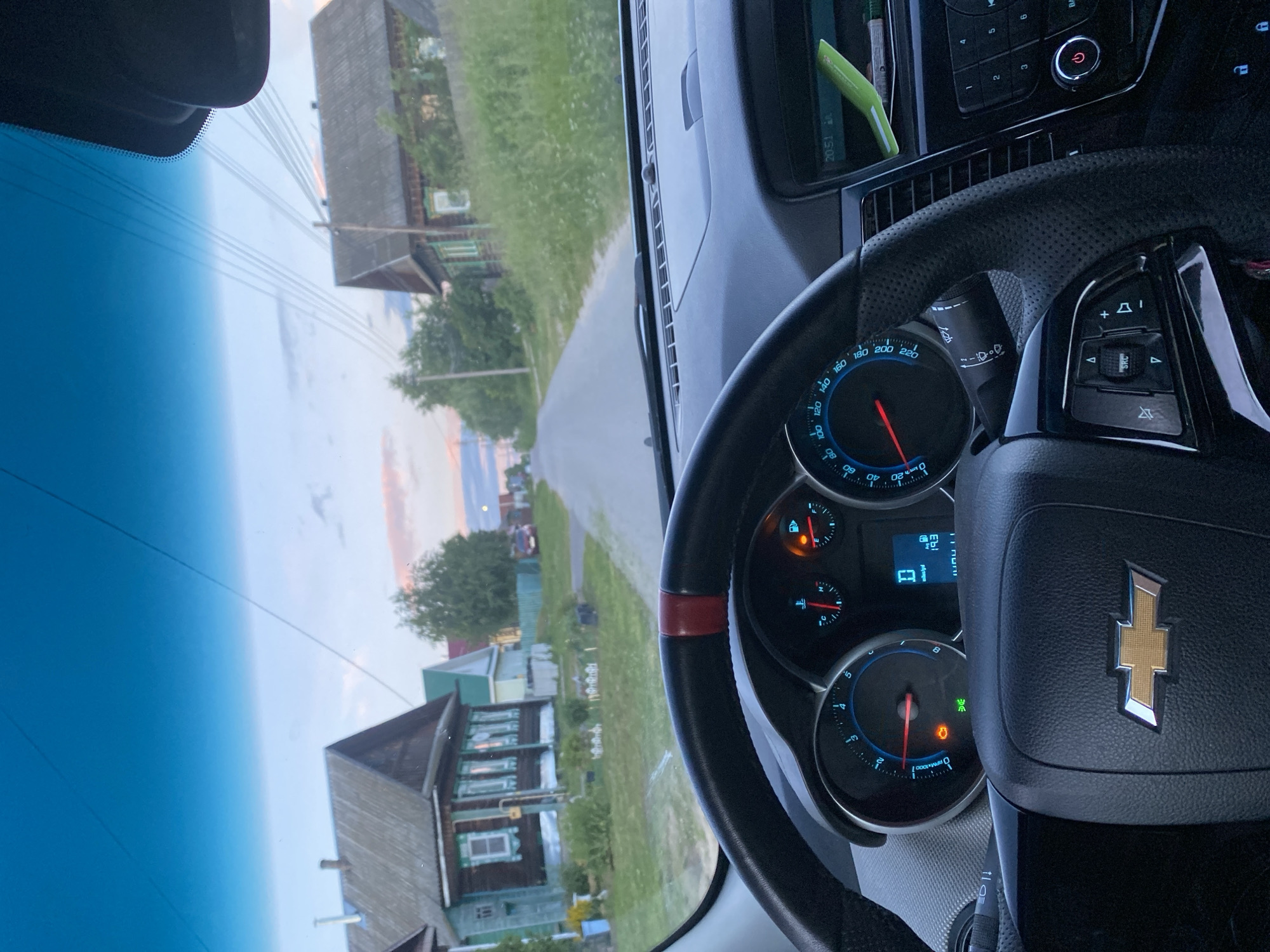The image shows a view from inside a car looking forward through the windshield. The car is driving on a paved road in what appears to be a rural or semi-rural area. On either side of the road are traditional wooden houses with green and brown exteriors and peaked roofs. Lush green grass and trees are visible. The sky is a gradient of blue with hints of pink and orange, suggesting it is either sunrise or sunset. The dashboard and steering wheel of the car are in the foreground, with the Chevrolet logo prominently displayed on the steering wheel. Dials and indicators on the dashboard are partially visible. The location is identified as Medvevo, Russia.
