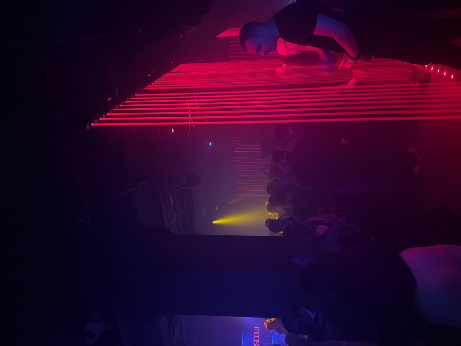 The image depicts an interior scene within a crowded nightclub or event venue in Moscow, Russia, identified by a discernible "MOSCOW" sign in blue illumination in the far-left background. The environment is characterized by low light and dynamic artificial illumination. A prominent feature on the right side is a vertical array of numerous parallel red light strips, approximately two meters in height, creating a luminous partition or backdrop. A male individual, wearing a dark short-sleeved shirt, stands on the far right in profile, facing left, with his right side illuminated by these red lights. The central and left-midground areas are densely populated with indistinct figures of individuals, primarily obscured by shadow and distance. A distinct yellow spotlight illuminates a portion of the crowd in the deeper background, creating a halo effect. A large, dark structural pillar is visible on the left, extending upwards. The ceiling is dark, with overhead fixtures, possibly speakers or additional lighting, visible. The overall scene conveys a typical nocturnal social gathering.