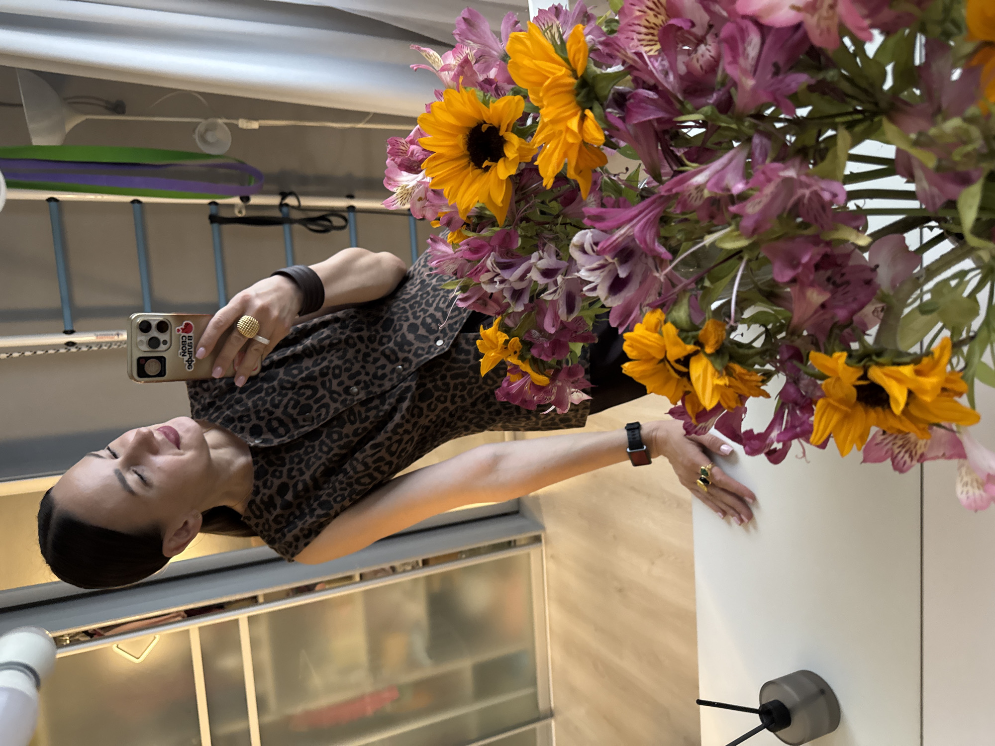 A woman with dark hair pulled back in a ponytail is taking a mirror selfie indoors. She is wearing a sleeveless, brown and black leopard-print top and black pants. She is also wearing a gold ring with a textured surface on her right index finger and a dark leather bracelet on her right wrist. In her right hand, she holds a gold smartphone. In the foreground, a bouquet of colorful flowers, including sunflowers and pink alstroemeria, is displayed on a white surface. To the left of the flowers, a dark grey reed diffuser is visible. The background shows a closet with glass doors, a ladder-like structure with colored bands, a floor lamp, and a window with curtains. The floor appears to be light-colored wood. The scene suggests an indoor residential setting. There is no indication of the location being the city of Sochi, Russia.