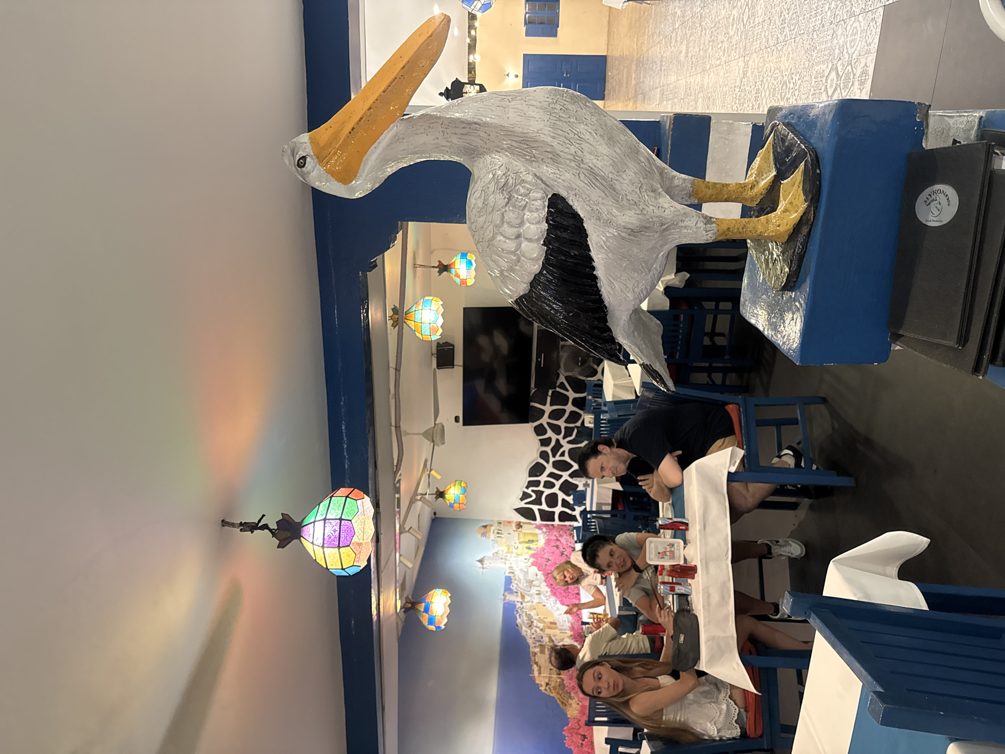 The image depicts the interior of a restaurant with a Mediterranean-inspired theme, likely located in North Kuta, Indonesia. In the foreground, a large statue of a pelican stands on a blue pedestal, its white body contrasting with black wing markings and a bright yellow beak and feet. The pelican's gaze is directed upwards and to the right.
The restaurant features blue and white striped pillars and blue wooden chairs with white cushions. Tables are covered with white tablecloths. Several patrons are seated at tables, engaged in dining and conversation. On the left, two women are seated at a table, one in a white dress and the other in a grey t-shirt. Another woman is visible further back, holding a drink. On the right, a man in a black t-shirt and khaki shorts is seated at a table, his arms crossed.
The walls are adorned with a mural depicting a white village with pink flowers and a blue sea. Colorful, stained-glass pendant lights hang from the ceiling, casting a vibrant, diffused light onto the white ceiling and walls. A large flat-screen television is mounted on a wall in the background. In the far background, blue shutters and a doorway suggest an exit or another section of the restaurant. The floor is tiled with a light-colored geometric pattern. Menus are stacked on a surface in front of the pelican statue.