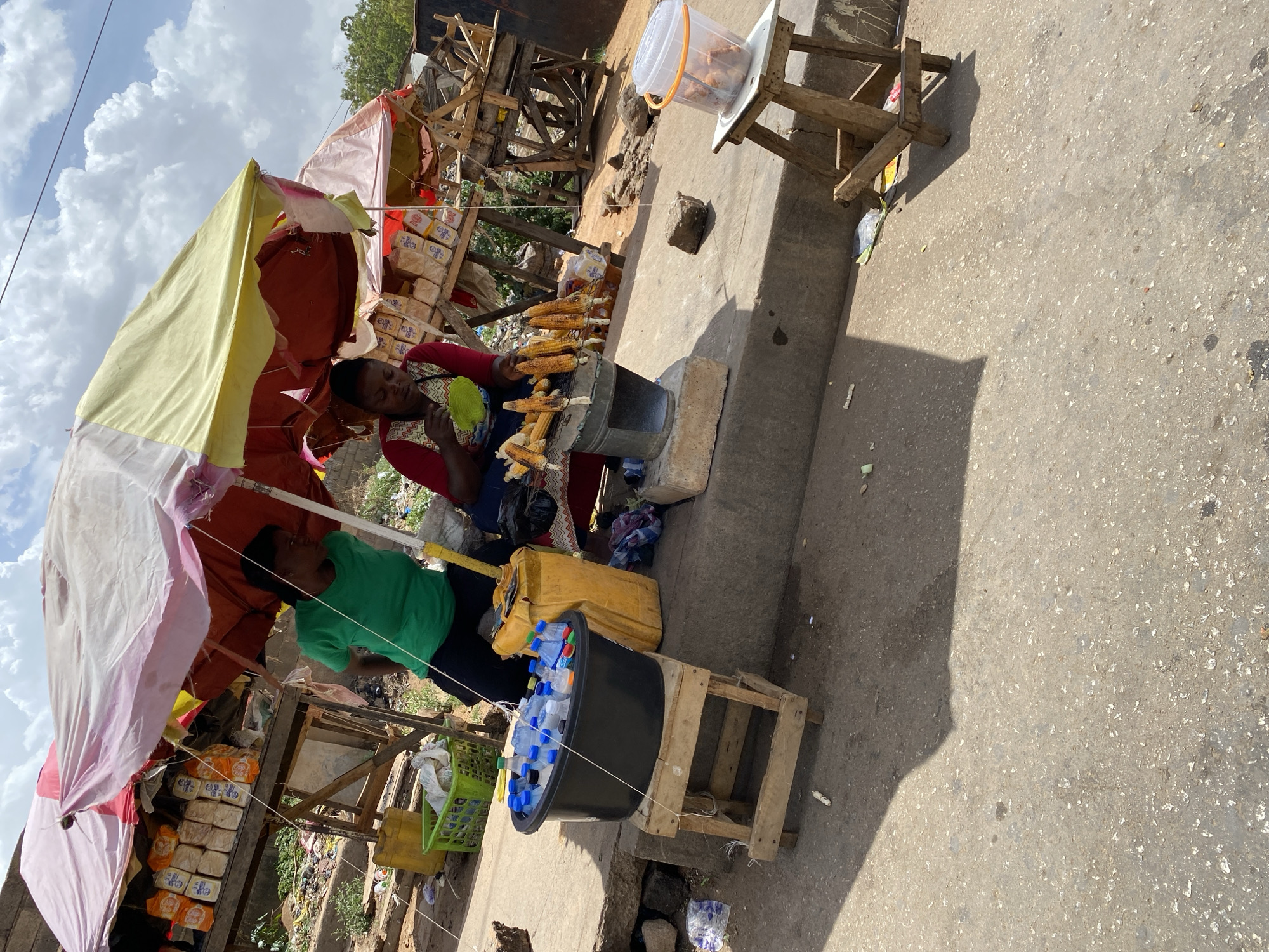 This outdoor image, taken in Bukuru, Nigeria, captures a bustling informal market scene under bright daylight with a partially cloudy sky, casting strong shadows. The perspective is slightly elevated, looking down into the market area.
The central activity features a woman, dressed in a red and patterned top, seated and actively fanning or grilling corn on the cob over a small, cylindrical metal stove. Several ears of roasted corn are displayed on the grill, suggesting she is a vendor preparing food. Beside her, another woman in a green t-shirt stands near the stall, possibly a customer or assistant.
The market stall is makeshift, consisting of various wooden structures. To the left of the woman in red, numerous loaves of packaged bread are stacked on a wooden stand. The packaging clearly shows text, including "FLOUR MILLS OF NIGERIA PLC" and "GOLDEN PENNY." An umbrella, with yellow, red, and pink fabric sections, provides shade over the stall.
Other items for sale or use are visible: a black tub filled with bottled water sits on a small wooden stand, and a yellow jerrycan is placed nearby. Another clear plastic bucket, possibly holding fried snacks or dough, rests on a smaller wooden stand further along the concrete path. The ground is a mix of concrete and dirt, with some debris indicative of an active market environment. In the background, more informal structures and vegetation are visible, suggesting a vibrant local trading area.