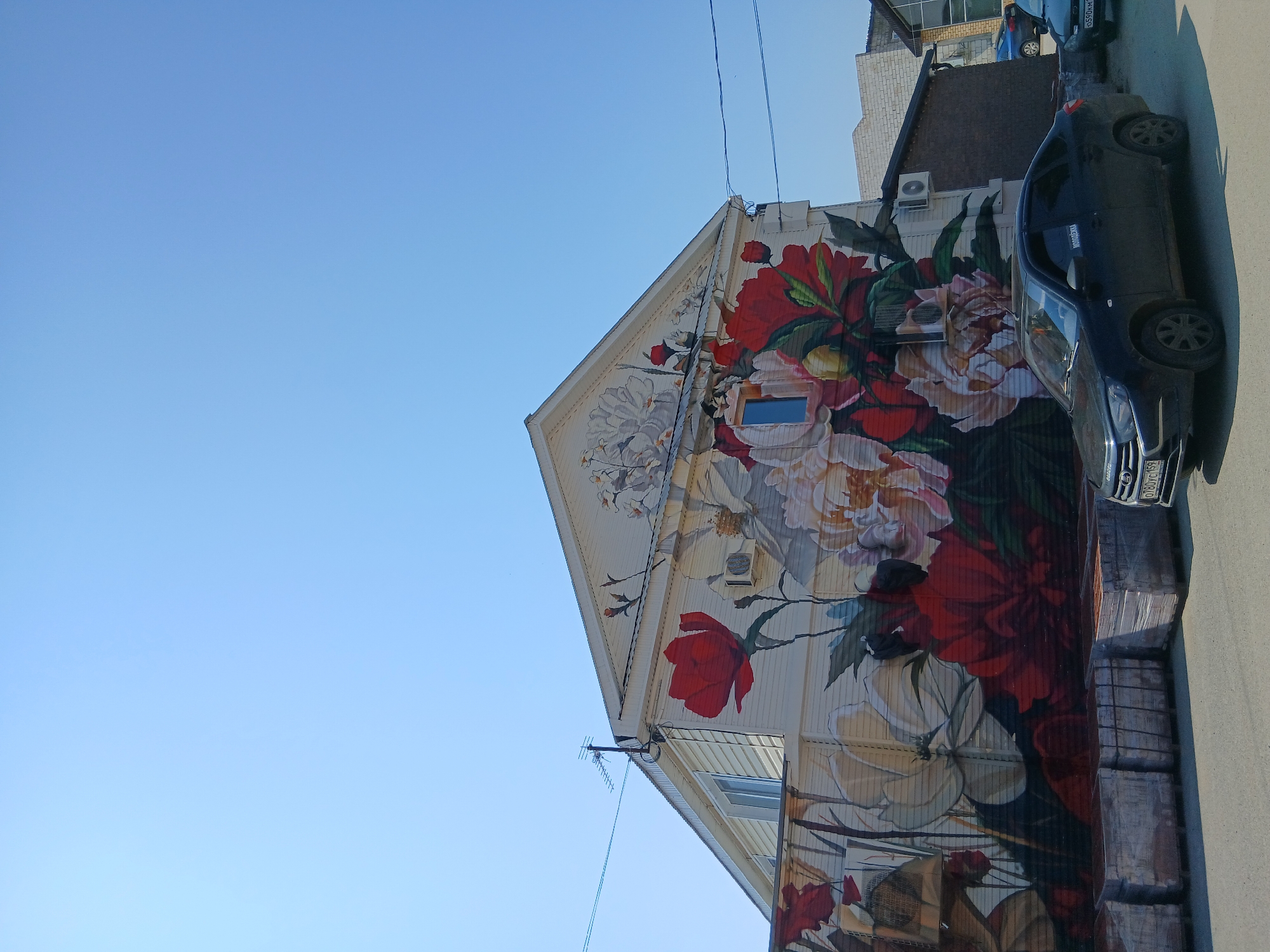 The image shows a building in the city of Osa, Russia, adorned with a large mural of peonies. The building has a light-colored siding and a triangular roof. A window with blue glass is visible on the facade. In front of the building, several pallets of what appear to be bricks are stacked on the pavement. Two cars are parked on the street in front of the building. One is a dark-colored sedan, and the other is partially visible and appears to be a similar color. The sky above is clear and blue. The scene suggests a sunny day in a commercial or residential area.