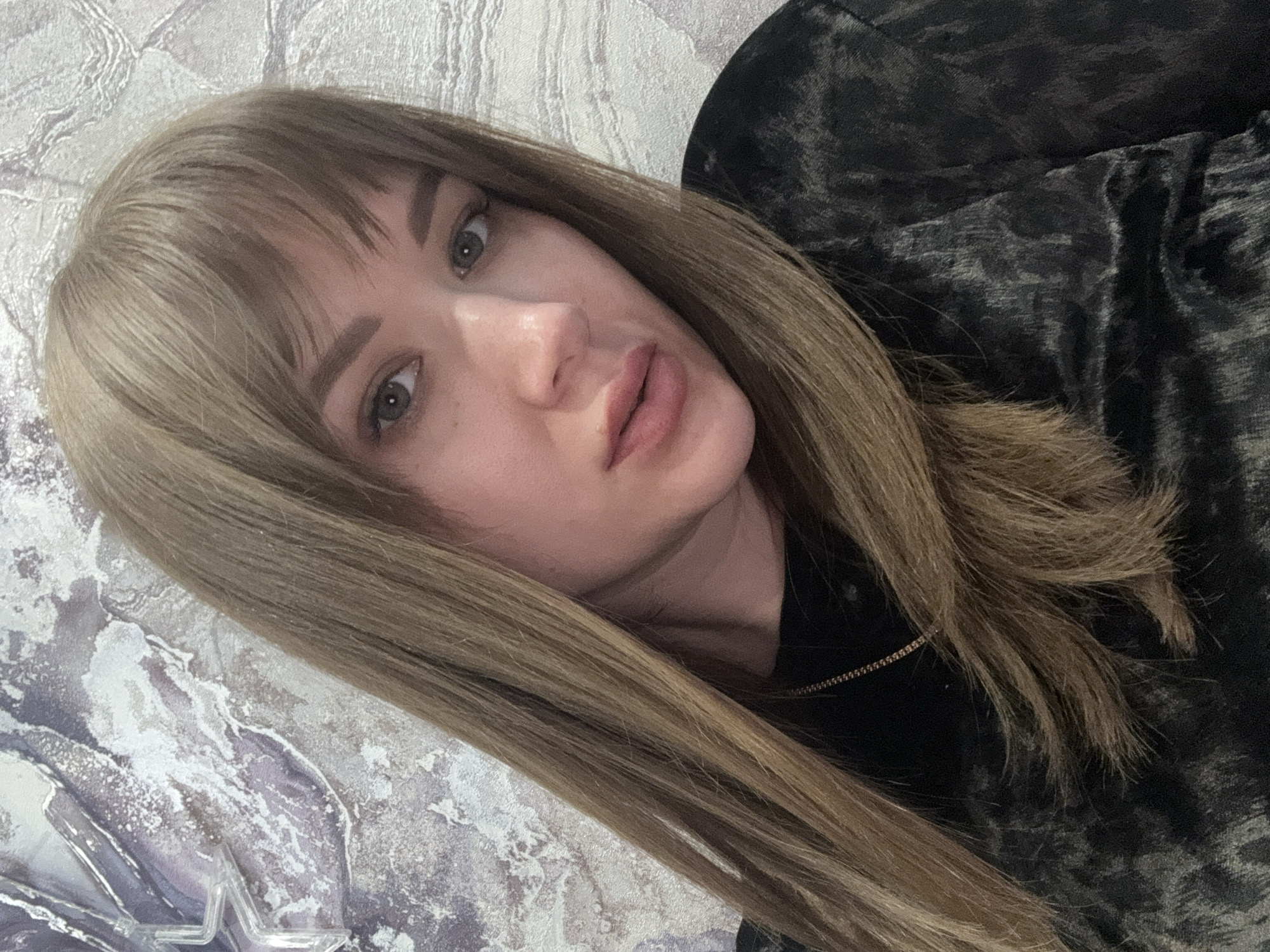 A woman with light brown hair and bangs is pictured indoors. Her hair is long and straight, falling towards the left side of the frame. She has fair skin, light-colored eyes, and pink lips. She is wearing a dark patterned top. The background behind her is a wall or surface with a marbled pattern in white and gray. Part of a small clear plastic object is visible on the lower left side. No specific location within Chaikovsky, Russia, is identifiable from the image.
