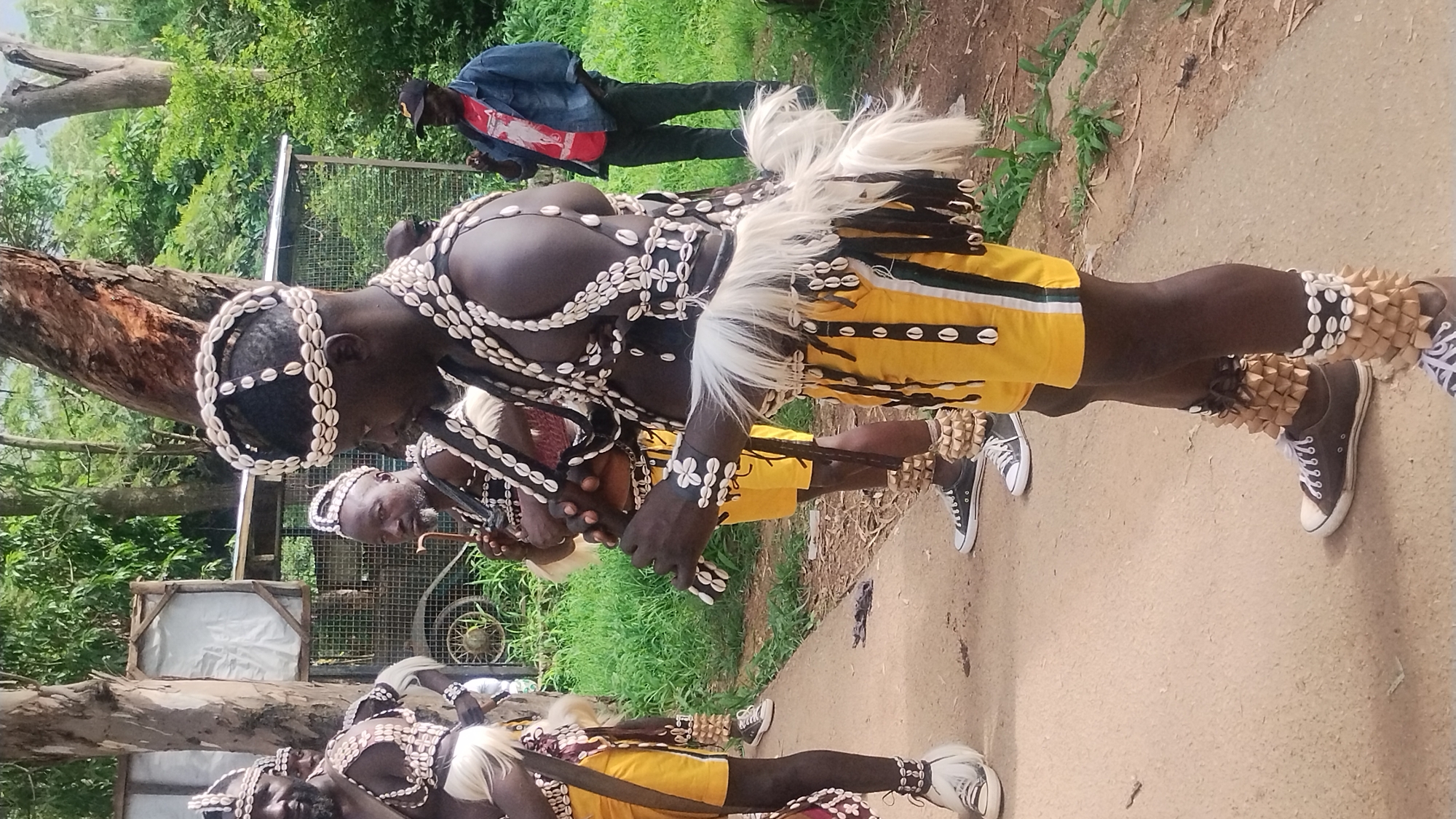 In Jos, Nigeria, a group of men in traditional attire stand on a dirt path surrounded by trees and greenery. The men are adorned with elaborate headdresses and chest decorations made of white shells. They wear yellow shorts with white and green stripes and beaded anklets. One man plays a flute-like instrument. In the background, a man in a denim jacket and a red shirt stands observing. A wire fence and what appears to be a weathered structure are visible behind the performers.