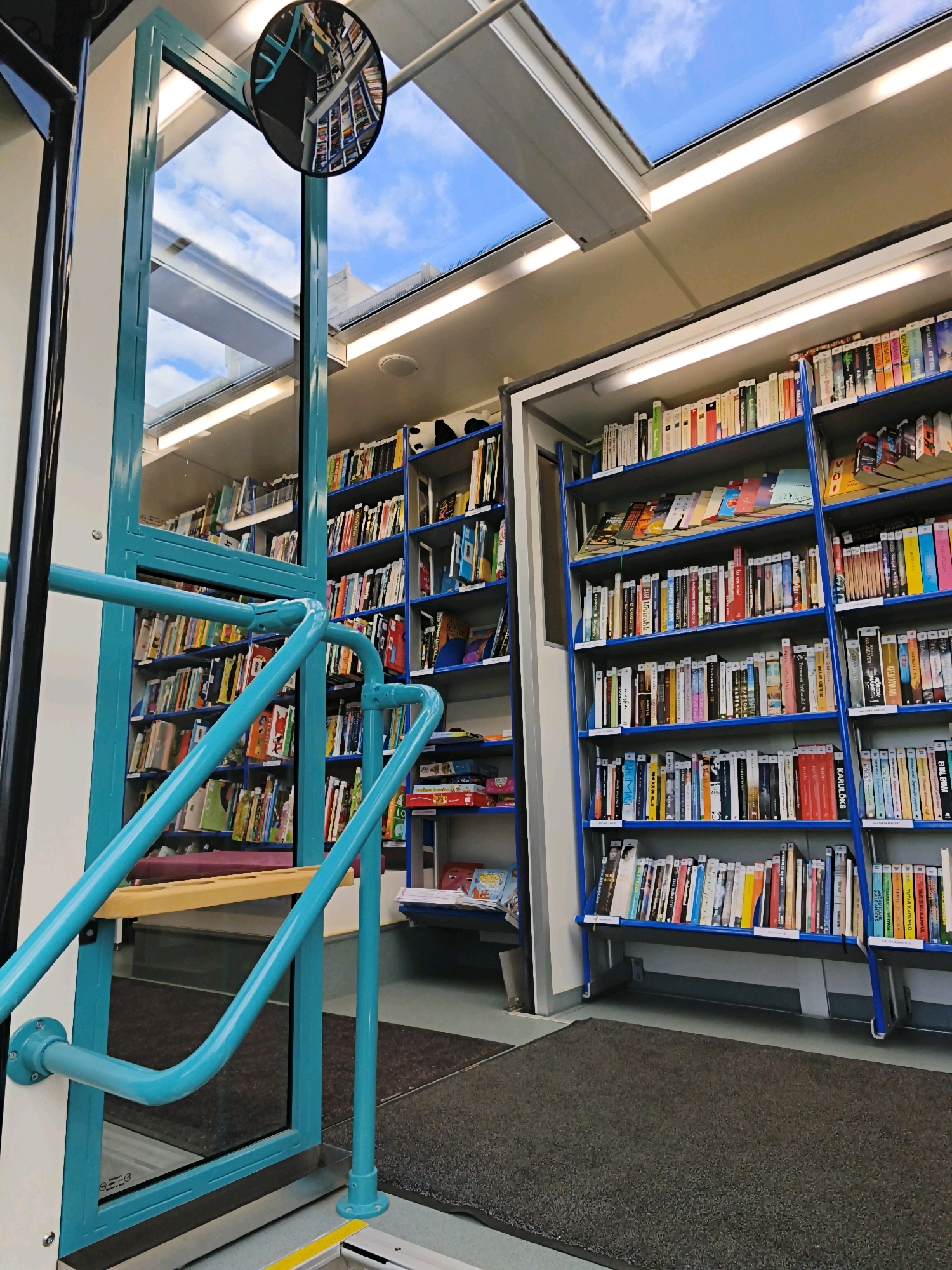 The image captures the interior of a mobile library, likely located in the city of Tallinn, Estonia, given the reflected text on the mirror which appears to be in Cyrillic script and suggests a location within Eastern Europe. The library is housed within a structure that features a large glass ceiling, allowing natural light to illuminate the space and offering a view of the blue sky with scattered clouds.
The main subjects are bookshelves packed with numerous books, arranged in neat rows across multiple levels. The shelves are a bright blue, contrasting with the lighter tones of the books and the environment. A prominent turquoise-colored handrail system is visible in the foreground, indicating a set of stairs or an elevated platform within the mobile library. This railing is robust and appears to be made of metal.
On the left side of the image, a vertical door or entrance is visible, also framed in the same turquoise color. A convex mirror is mounted above this entrance, reflecting a portion of the interior bookshelves and the sky. The floor is covered with dark gray carpeting or mats.
The overall impression is that of a well-organized, accessible, and modern mobile library designed to bring books to the public. There are no people visible in the scene. The scene is clean and orderly, suggesting recent use or preparation for operation. The lighting is a mix of natural light from the ceiling and artificial light from unseen fixtures integrated into the ceiling structure.