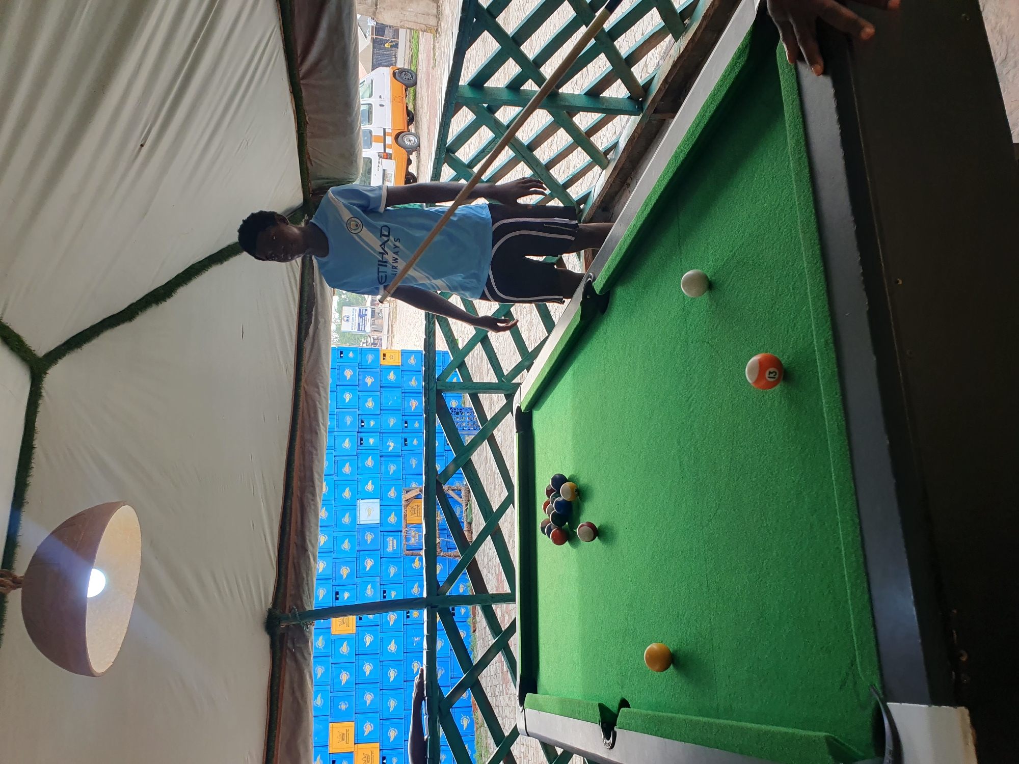 A young man stands beside a green billiard table, holding a cue stick in a partially enclosed area in Lokoja, Nigeria. The individual is wearing a light blue soccer jersey with "Etihad Airways" printed on the front, along with dark shorts with light-colored stripes. He appears to be taking aim at the pool balls. The billiard table holds a set of racked billiard balls, a white cue ball, and two solitary billiard balls. Behind him, a wall is constructed of stacked blue plastic crates. The area is partially covered by a tent-like structure with a light fixture hanging inside. Outside the enclosure, an ambulance is parked on a street.