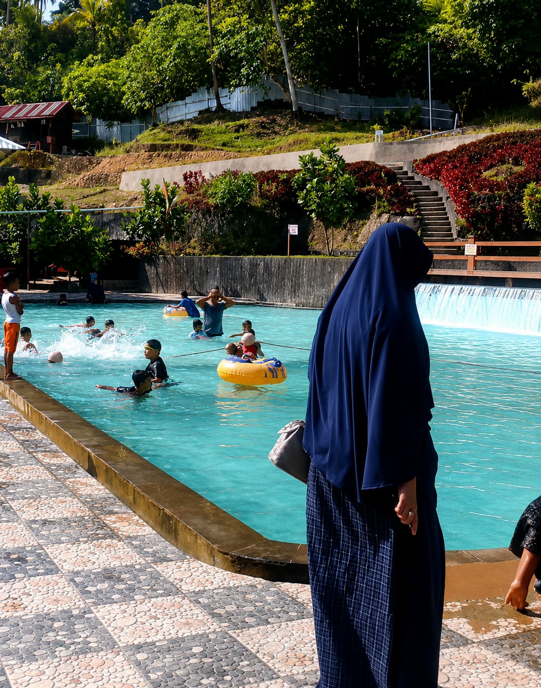 The image depicts a lively scene at a water park in Silaiyajae, Indonesia. The focal point is a large, turquoise swimming pool filled with a diverse group of people, primarily children, enjoying various water activities. In the foreground, a woman dressed in a dark blue hijab and a plaid skirt stands facing away from the camera, holding a light gray purse. Her hand, adorned with a ring, is visible at her side. The pool itself is surrounded by a patterned tiled walkway and a brown-edged pool border.
Several children are actively engaged in the water. Some are swimming, while others are utilizing inflatable rings. One child is seen floating in a yellow inner tube. In the background, a man in a dark blue shirt is partially submerged in the water, with his arms raised. Further back, other individuals can be seen swimming and playing in the pool.
The environment surrounding the pool suggests a lush, tropical setting. Dense green trees and vegetation cover the hillside behind the water park. A small, red-roofed building is nestled on the hillside, and a set of concrete stairs leads up the embankment. A metal fence runs along the top of the hill. The water park appears to be built into the natural landscape, with terraced areas and strategically placed foliage. The overall atmosphere is one of recreational enjoyment on a sunny day.