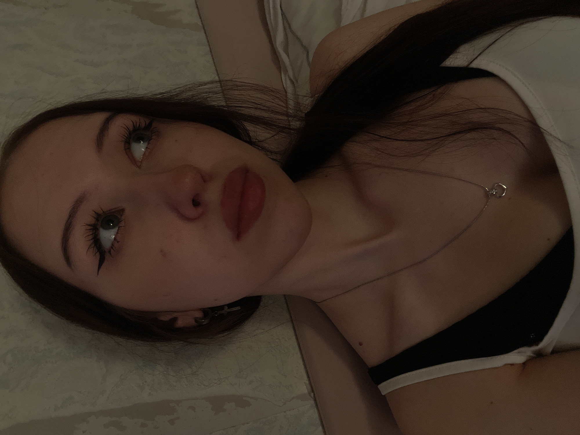 The image shows a close-up, eye-level shot of a young woman lying down. Her head is tilted to her right, and her eyes are looking up and to her right. She has dark hair, green eyes, and is wearing a black and white top. A silver necklace with a heart pendant is visible around her neck. The background appears to be a bedroom with textured wallpaper and bedding. The lighting is dim, casting shadows on her face and neck. The specific location in Votkinsk, Russia, cannot be determined from the visual information provided in the image.