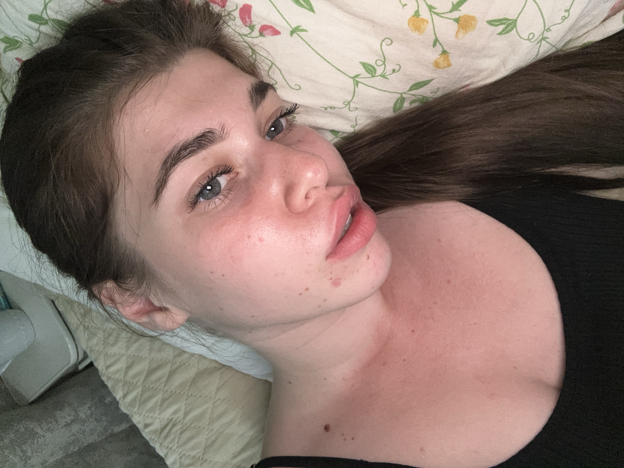 The image shows a close-up, overhead shot of a woman lying on a bed. She has long, dark brown hair, blue eyes, and is wearing a black top. The woman's face is tilted, and she is looking towards the camera. The bed appears to be covered with a light-colored, patterned duvet. The background is blurred and includes what appears to be a headboard and a bedside table with an electronic device. The lighting suggests an indoor setting, possibly a bedroom. The image does not contain any specific landmarks or contextual information that would allow for identification of the location as Мещёры, Russia.