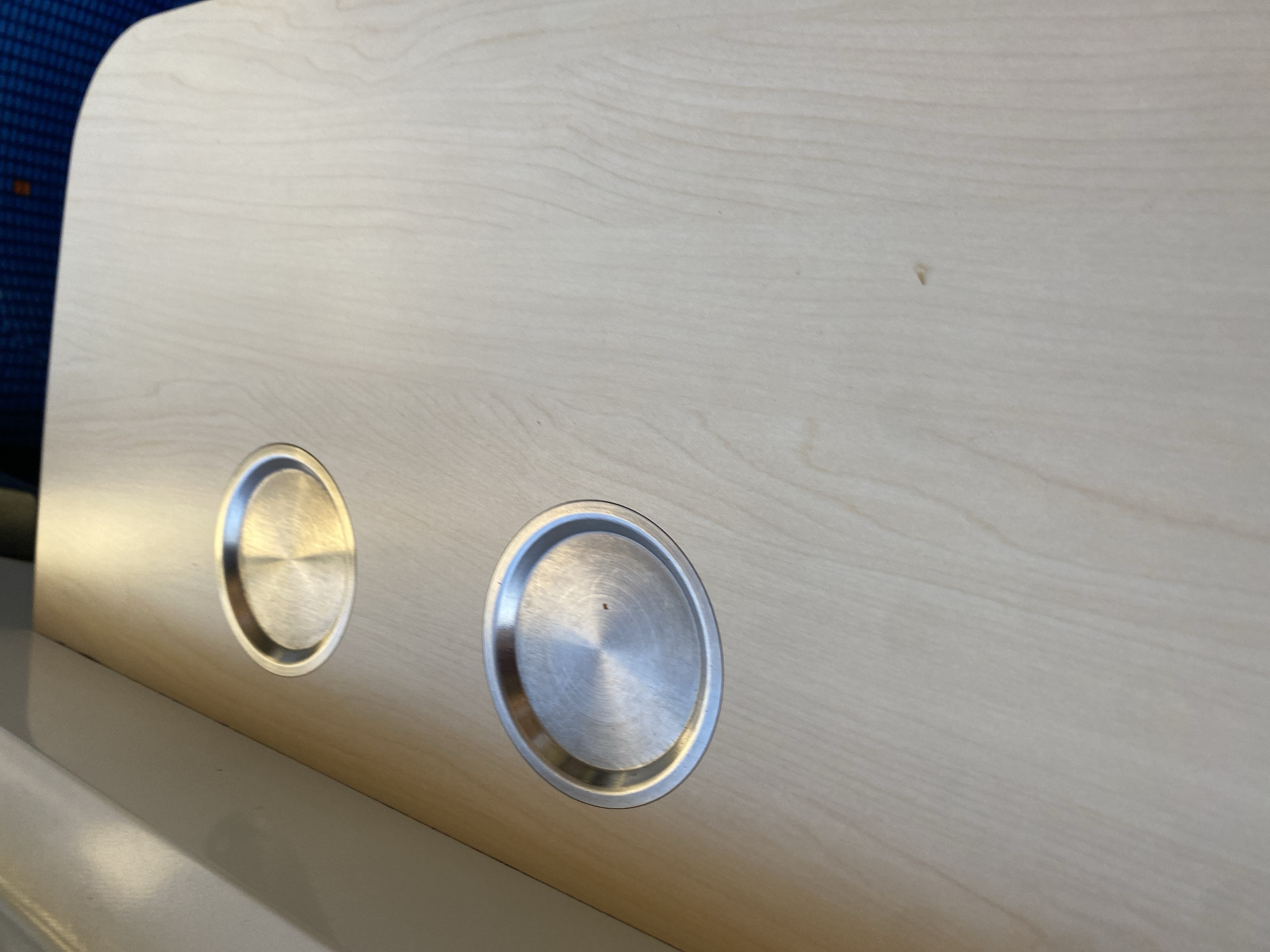 The image shows two circular, brushed metal cup holders embedded in a light-colored wooden surface. The cup holders are positioned at different distances from the viewer, with one slightly above and to the right of the other. The wooden surface has a visible grain pattern. The scene appears to be indoors, possibly on a table or desk. There is no indication of the location in České Zlatníky, Czechia.