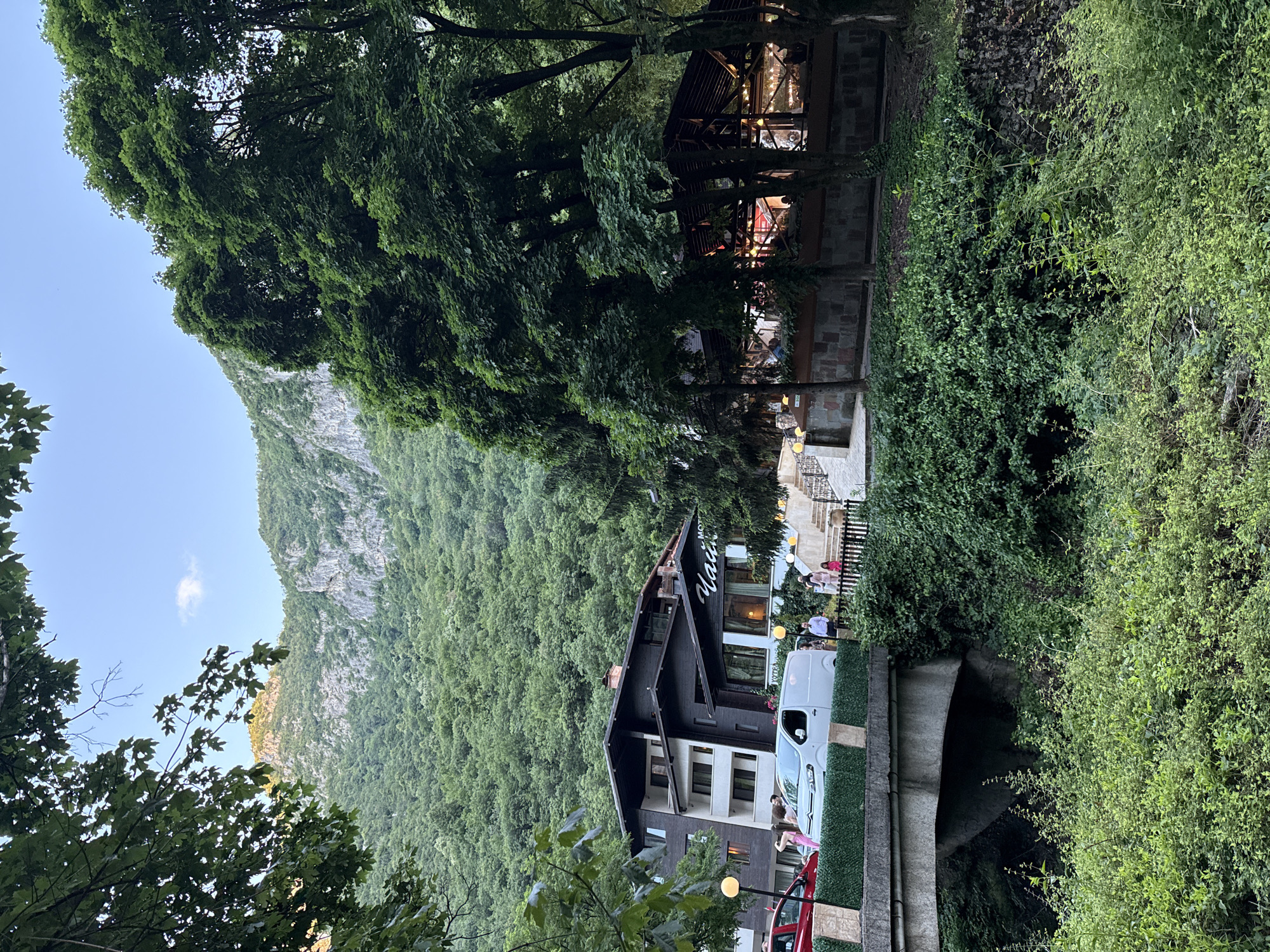 This image captures a scenic view of a hotel and restaurant complex nestled at the foot of a lush, tree-covered mountain range, likely in Vratsa, Bulgaria. The hotel, a multi-story building with a dark facade and white accents, is situated to the left, with several vehicles, including a white van and a red car, parked in front. A stone bridge crosses a small waterway in the foreground, with dense green foliage lining its banks and the area around the hotel. To the right of the hotel, an open-air restaurant with a wooden roof and stone base is illuminated, with patrons visible inside and around the structure. People are seen walking on pathways and stairs leading up to the hotel and restaurant. The sky above is clear with a hint of blue and a single white cloud, and the mountain peaks are illuminated by soft sunlight. The overall atmosphere suggests a tranquil, nature-infused retreat.