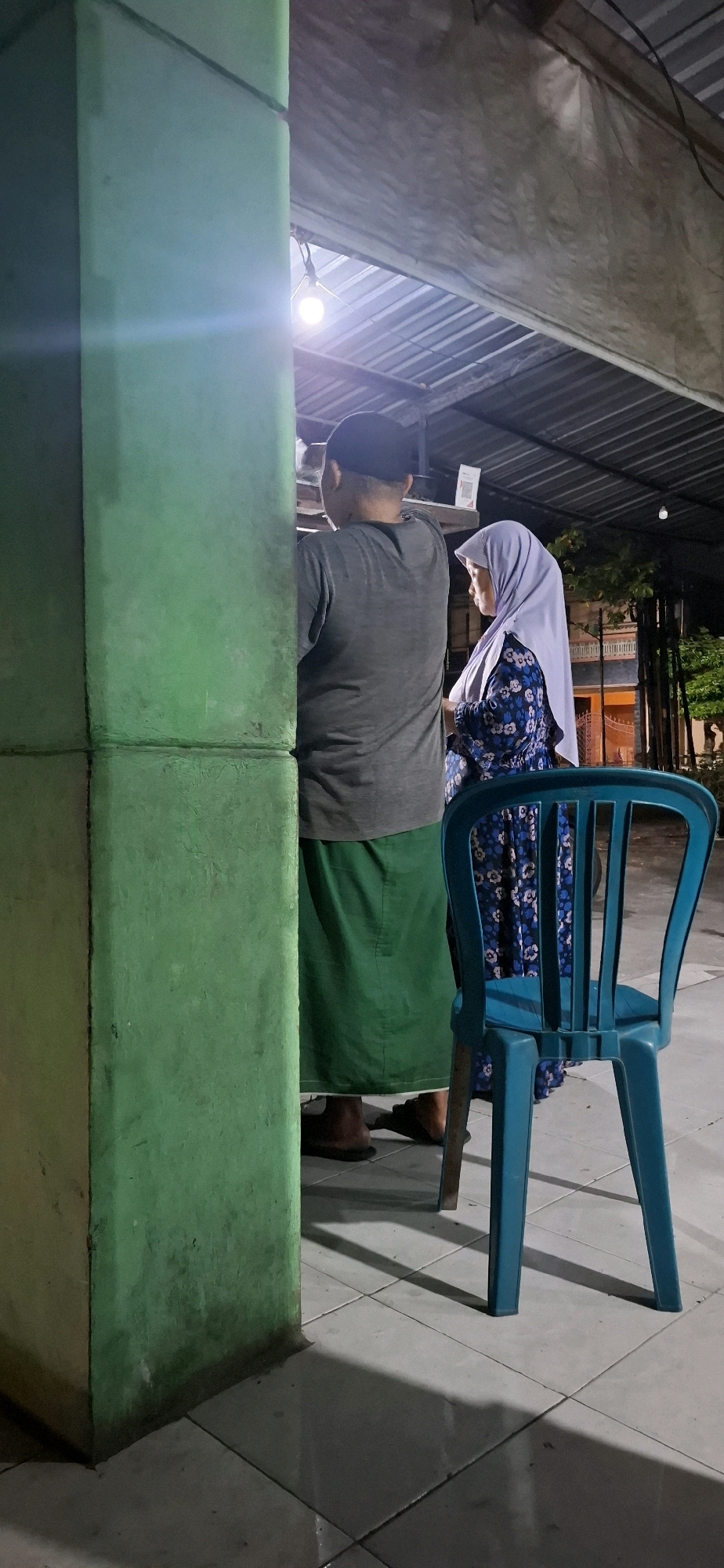 In Desa Gagaksipat, Indonesia, two individuals, a man and a woman, are standing near a stall. The man, wearing a grey t-shirt, a black cap, and a green sarong, is facing away from the camera. The woman, in a light purple hijab and a blue floral dress, is standing to his right and facing towards him. Behind them, a bright light bulb illuminates the area under a corrugated metal roof, which is supported by a green pillar on the left. In the foreground, a blue plastic chair is positioned to the right of the frame. The ground is tiled with square grey tiles, and shadows are cast by the objects and people present. The setting appears to be outdoors at dusk or night.