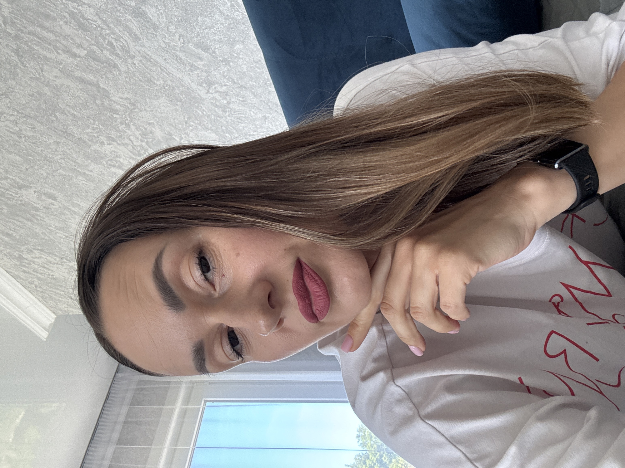 A young woman is depicted in a close-up portrait, originally oriented sideways but described as upright. She has long, light brown hair that cascades over her left shoulder and arm. She is wearing a white t-shirt adorned with red, stylized, and illegible text. Her left hand rests gently against her chin or cheek, showcasing light pink nail polish on her fingers. A black digital watch is visible on her left wrist. Her lips are accentuated with a dark pink or mauve lipstick, and her eyes are directed towards the camera with a neutral, contemplative expression.
The setting appears to be indoors during daytime, suggested by the bright natural light. The background includes a gray, textured surface, possibly a wall or ceiling. A dark blue sofa or cushion is partially visible behind her head. A window below offers a view of a clear blue sky and lush green foliage, indicating sunny weather. The provided context for the location is Sochi, Russia.