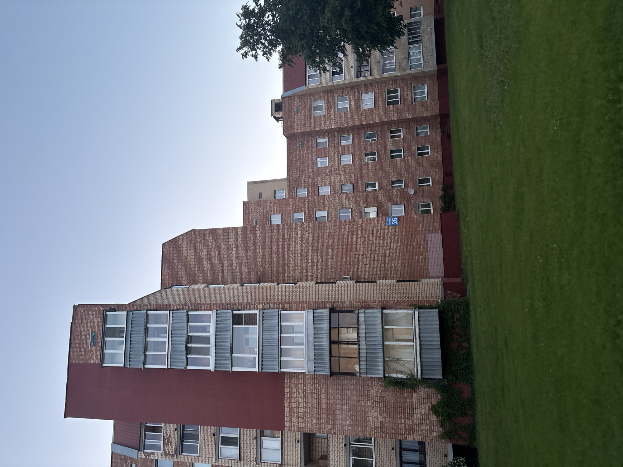 The image depicts a multi-story brick building with a combination of brick and modern window design elements, located in Minsk, Belarus. The building faces a green lawn area. The sky above is pale blue. The building facade is predominantly red brick with sections of red painted material. The windows range from traditional rectangular windows to modern, larger window units with metal frames. Vegetation is visible along some of the building's lower levels. A tree's crown partially obscures the building's upper levels. A blue address plate with the number "26" is attached to the building.