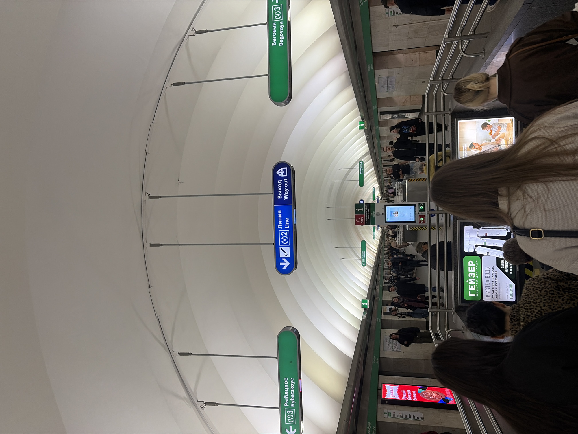 This is a view inside a metro station in Saint Petersburg, Russia. The photo is taken at an angle looking upward to the ceiling, which has a curved design with rows of lights. There are numerous people on the platform and on the escalators. Various signs are visible, including signs for "Begovaya" (Line 3), "Rybatskoye" (Line 3), "Line 2", and "Way Out". Advertisements are posted along the platform walls. The atmosphere appears crowded.